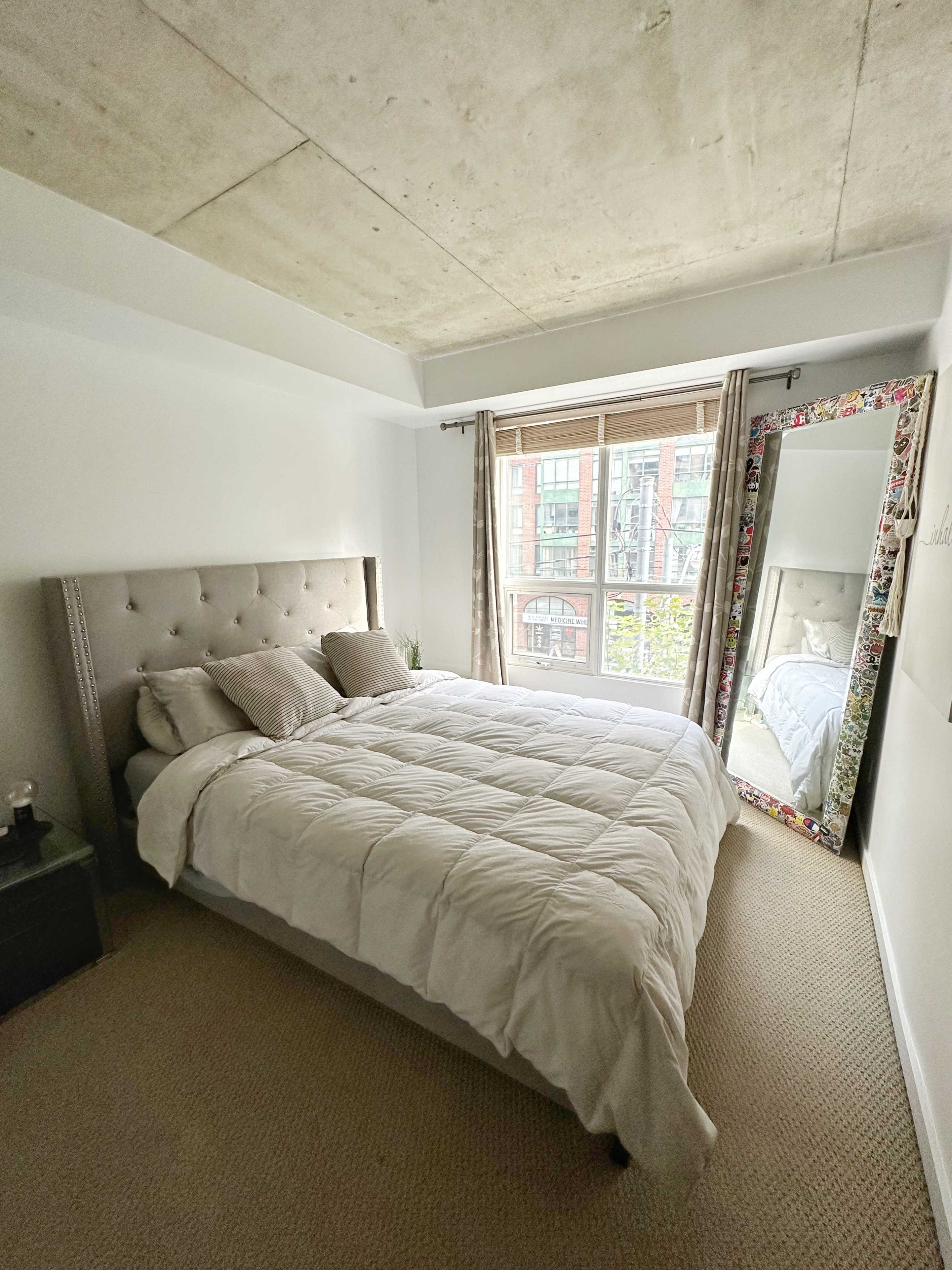 A tidy bedroom featuring a bed with a white comforter, a bedside table, and a large mirror next to a window with natural light.