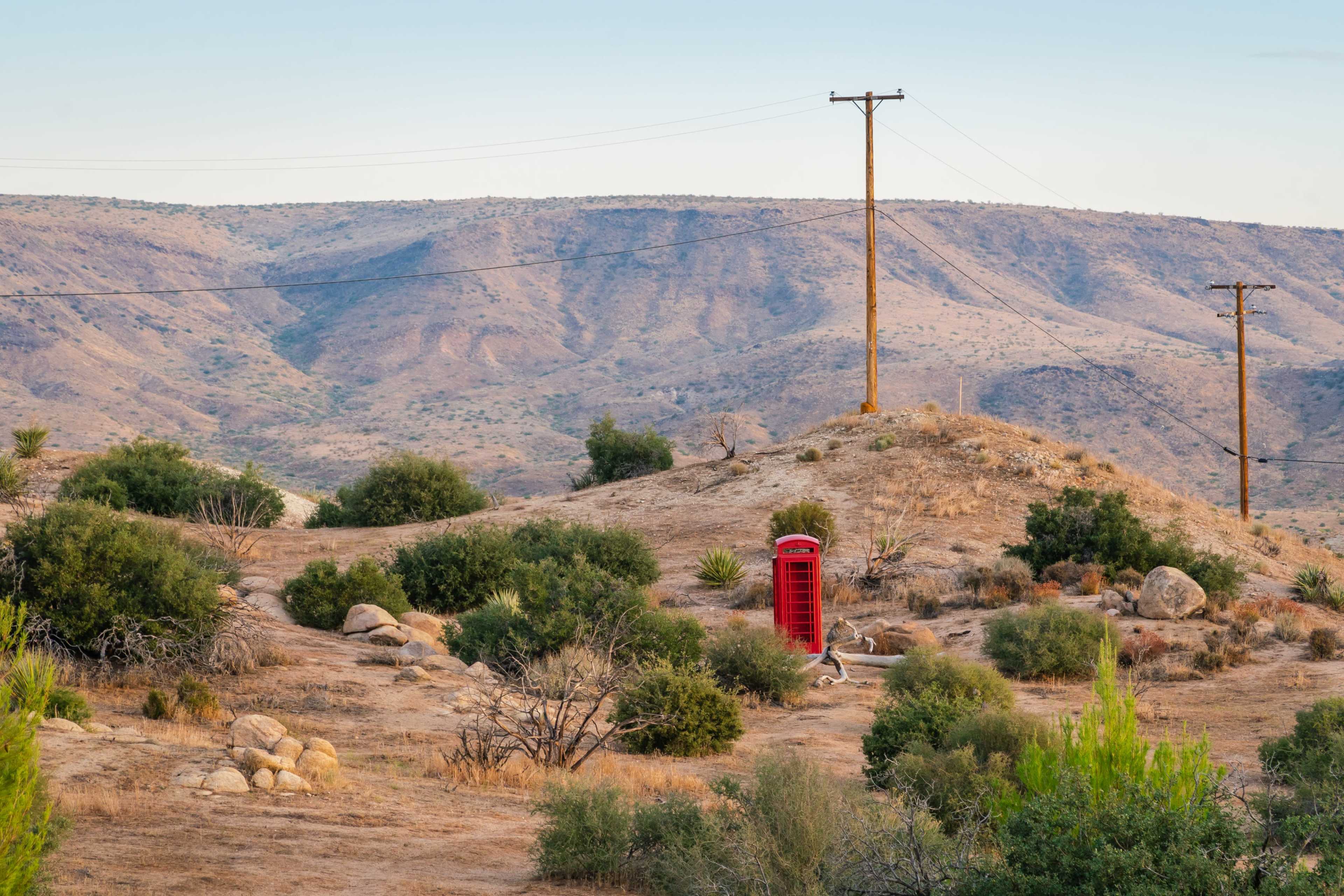 A red telephone booth stands alone on a hillside surrounded by dry vegetation and rolling mountains.