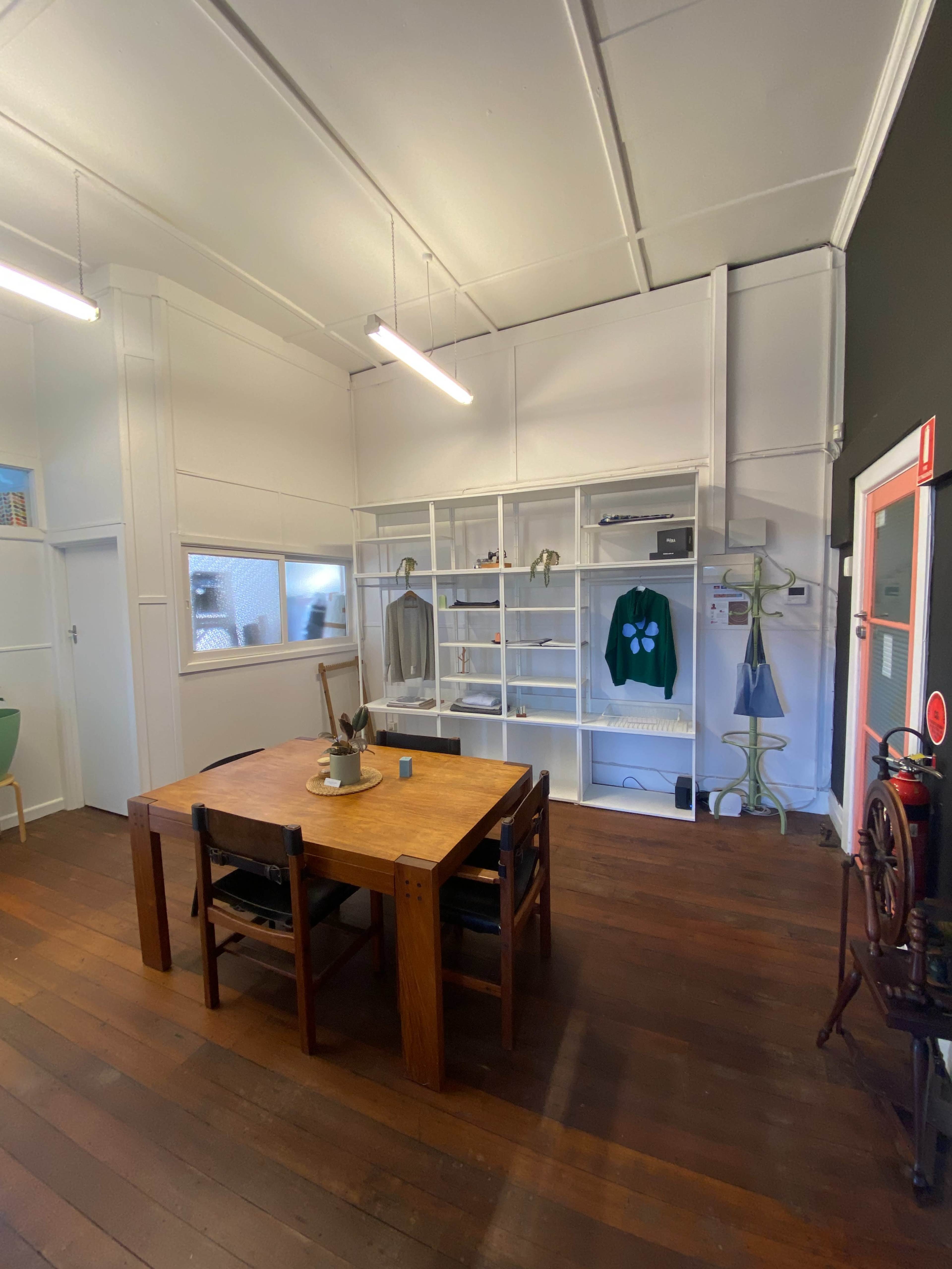 The image shows a room with wooden flooring, a table with chairs in the foreground, and a wall-mounted shelf displaying clothing and accessories.