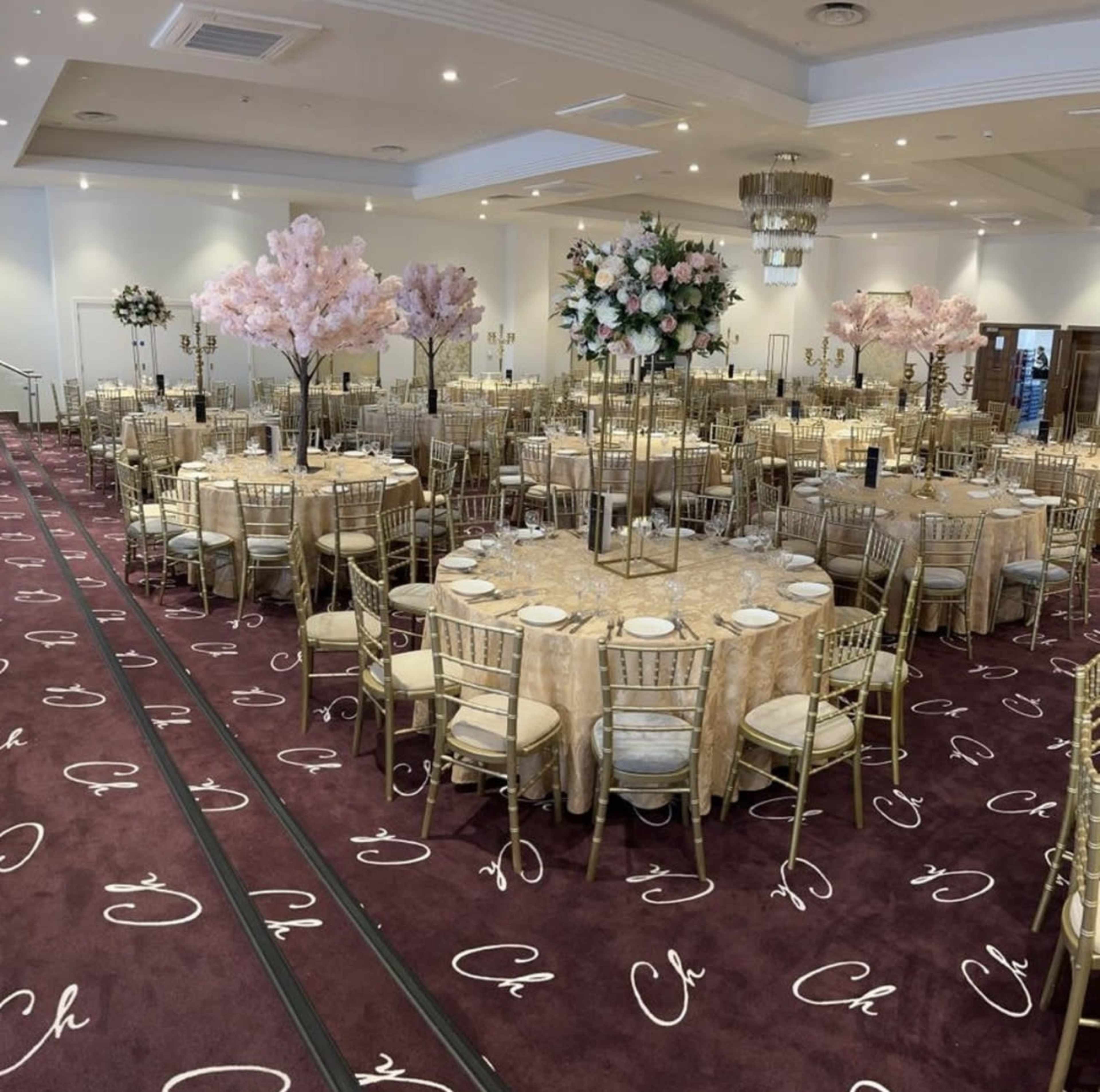 The image shows a banquet hall set up with round tables adorned with beige tablecloths and floral centerpieces, surrounded by gold chairs, with pink cherry blossom trees positioned at various points in the room.