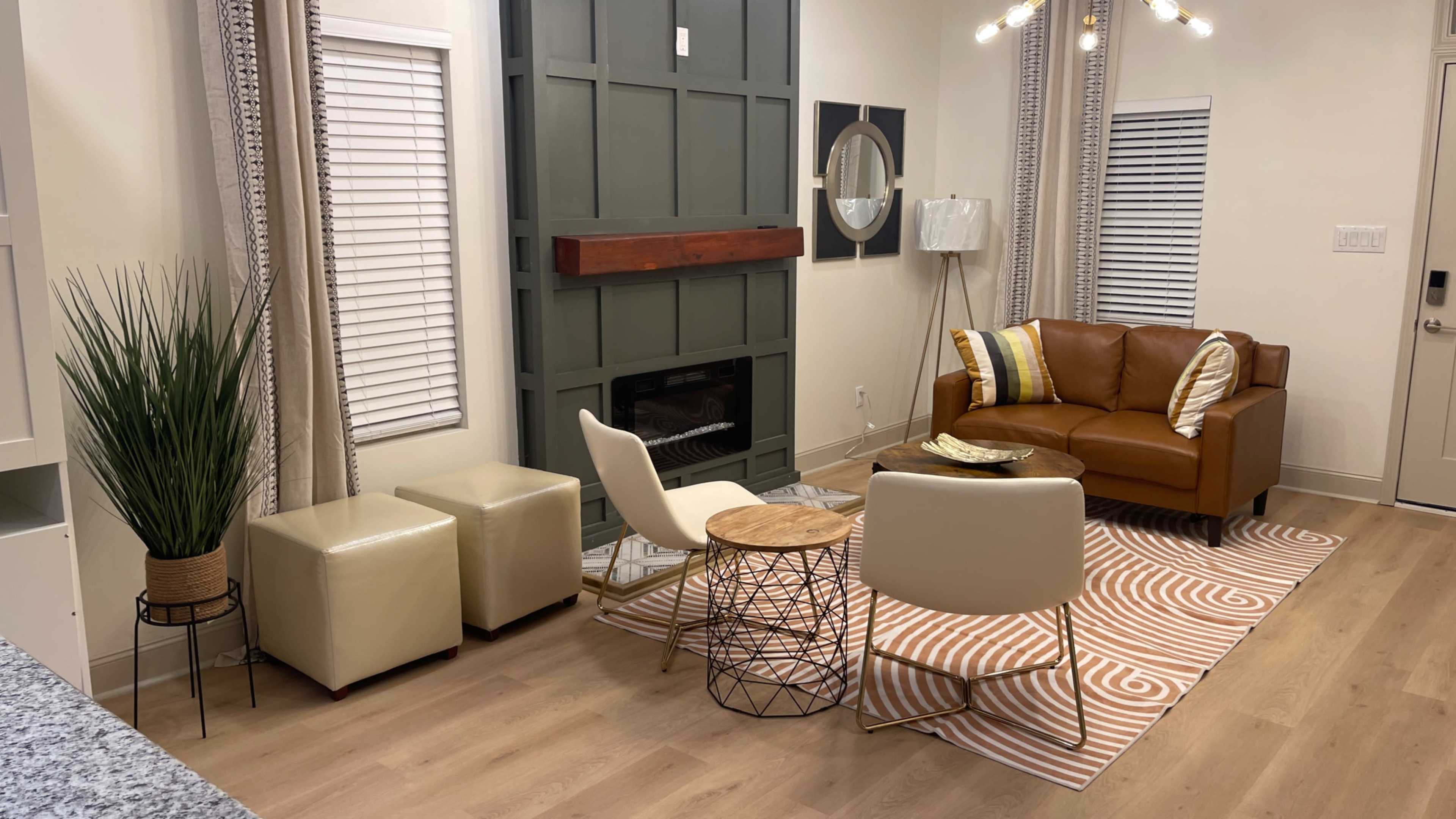 A modern living room with a brown leather sofa, a round wooden coffee table, and cream-colored accent chairs arranged around a patterned area rug.