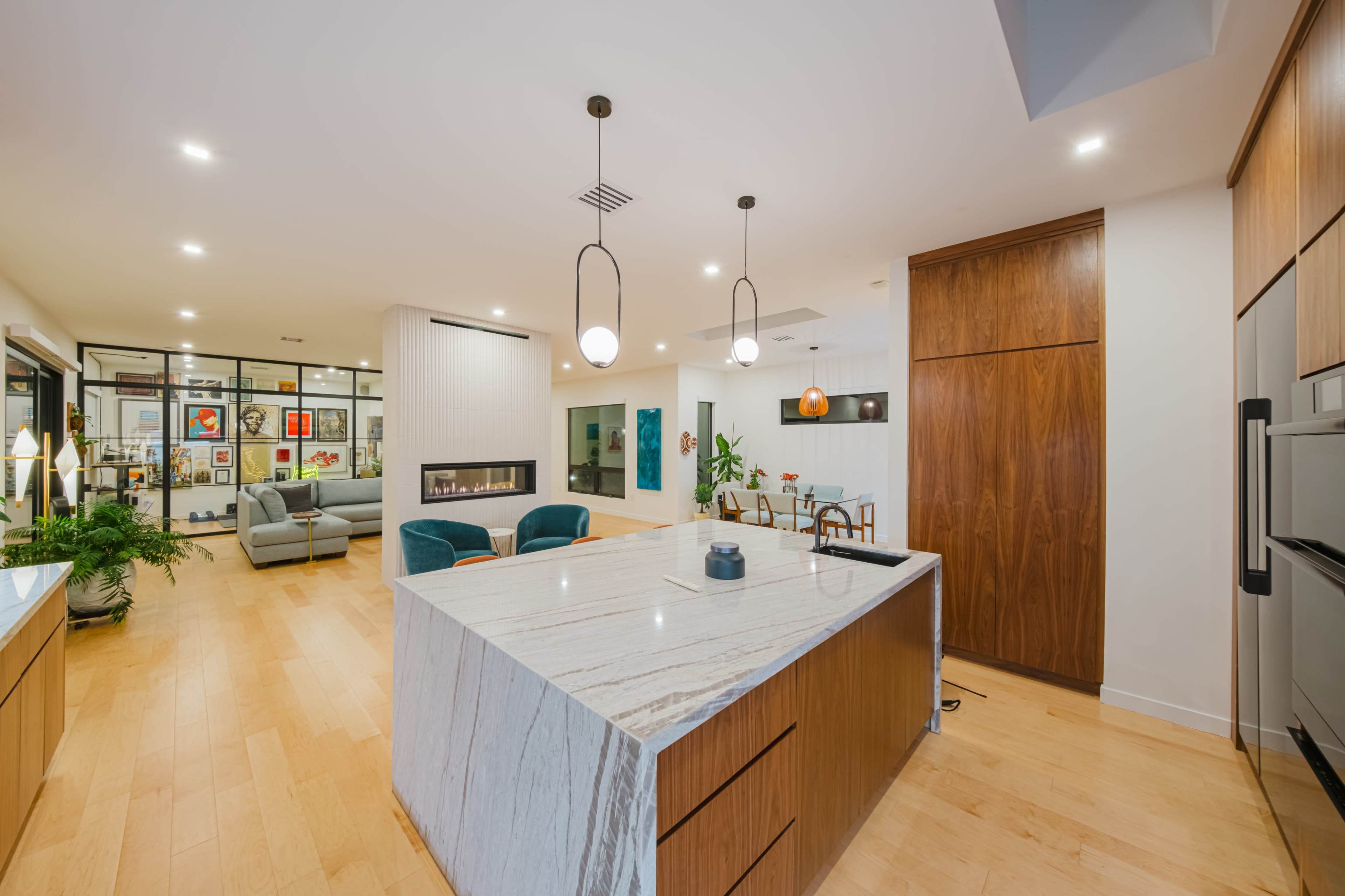 The image shows a modern open-concept kitchen and living space, featuring a large marble island and wooden cabinetry, with comfortable seating and artwork displayed on the walls.