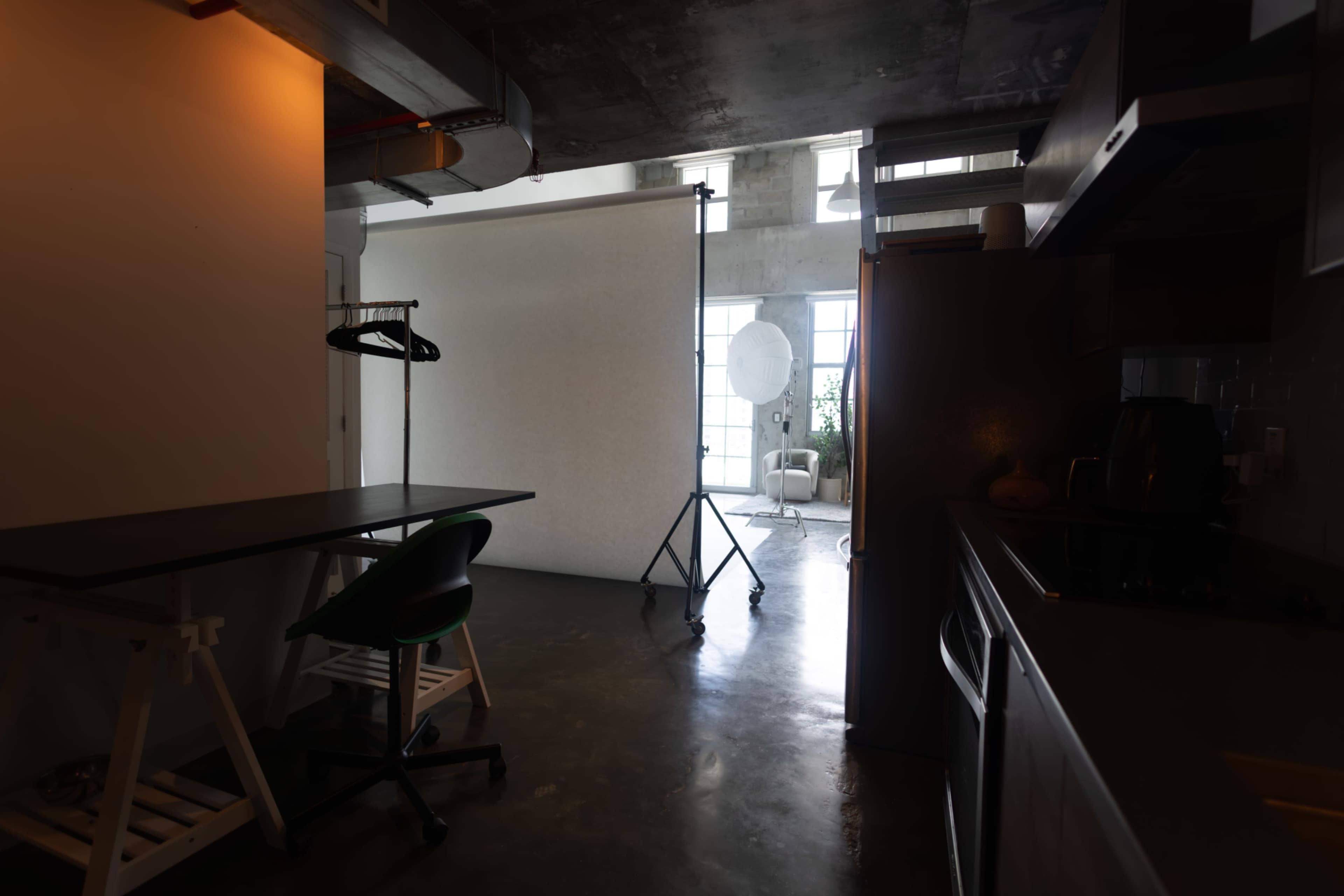 A modern studio space features a kitchen area and a backdrop setup with natural light illuminating the scene through large windows.