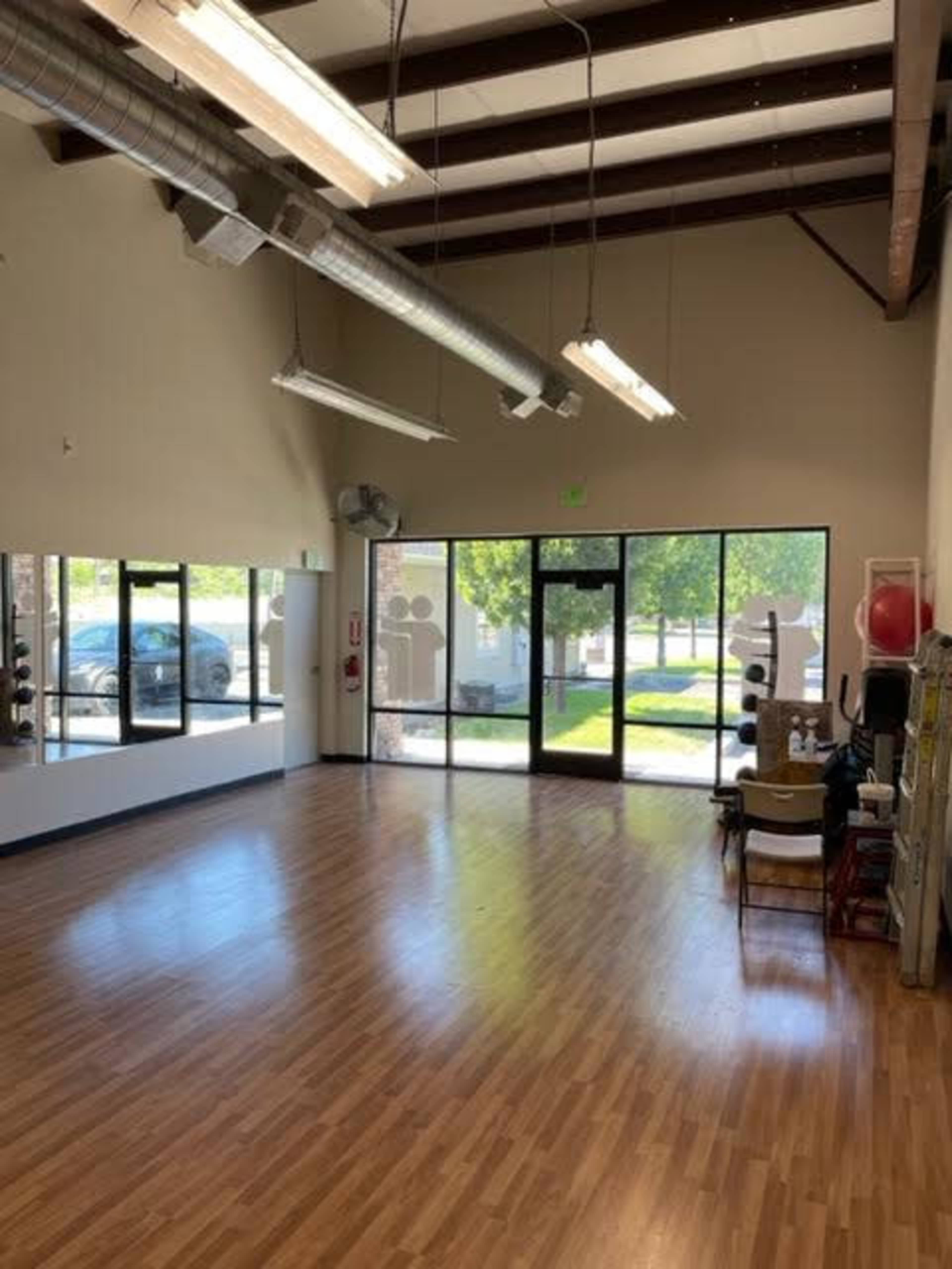 A spacious indoor area with wooden flooring, large windows allowing natural light, and a few chairs arranged near the walls.