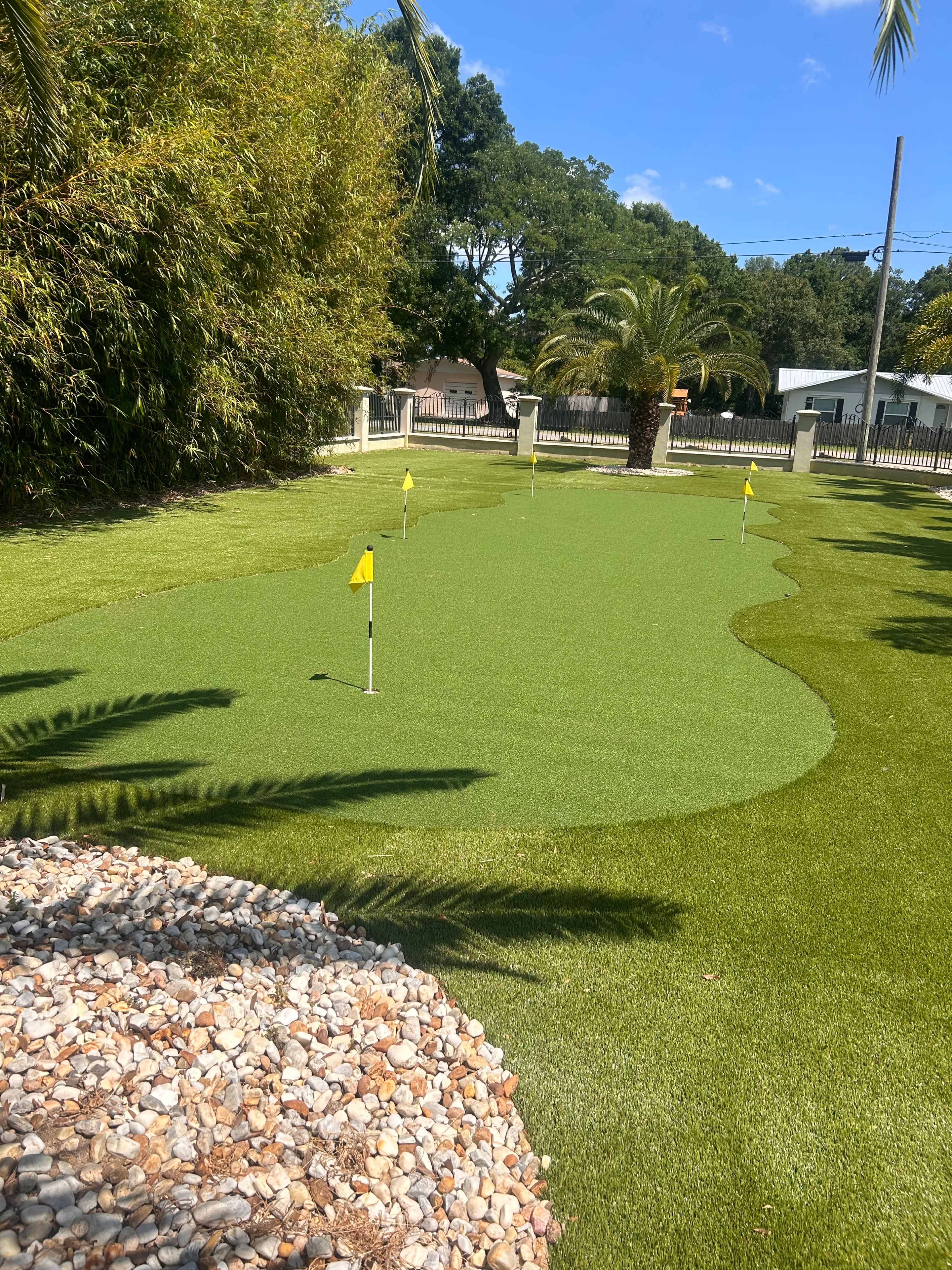 A green artificial putting surface with several holes marked by yellow flags, surrounded by lush landscaping and palm trees.