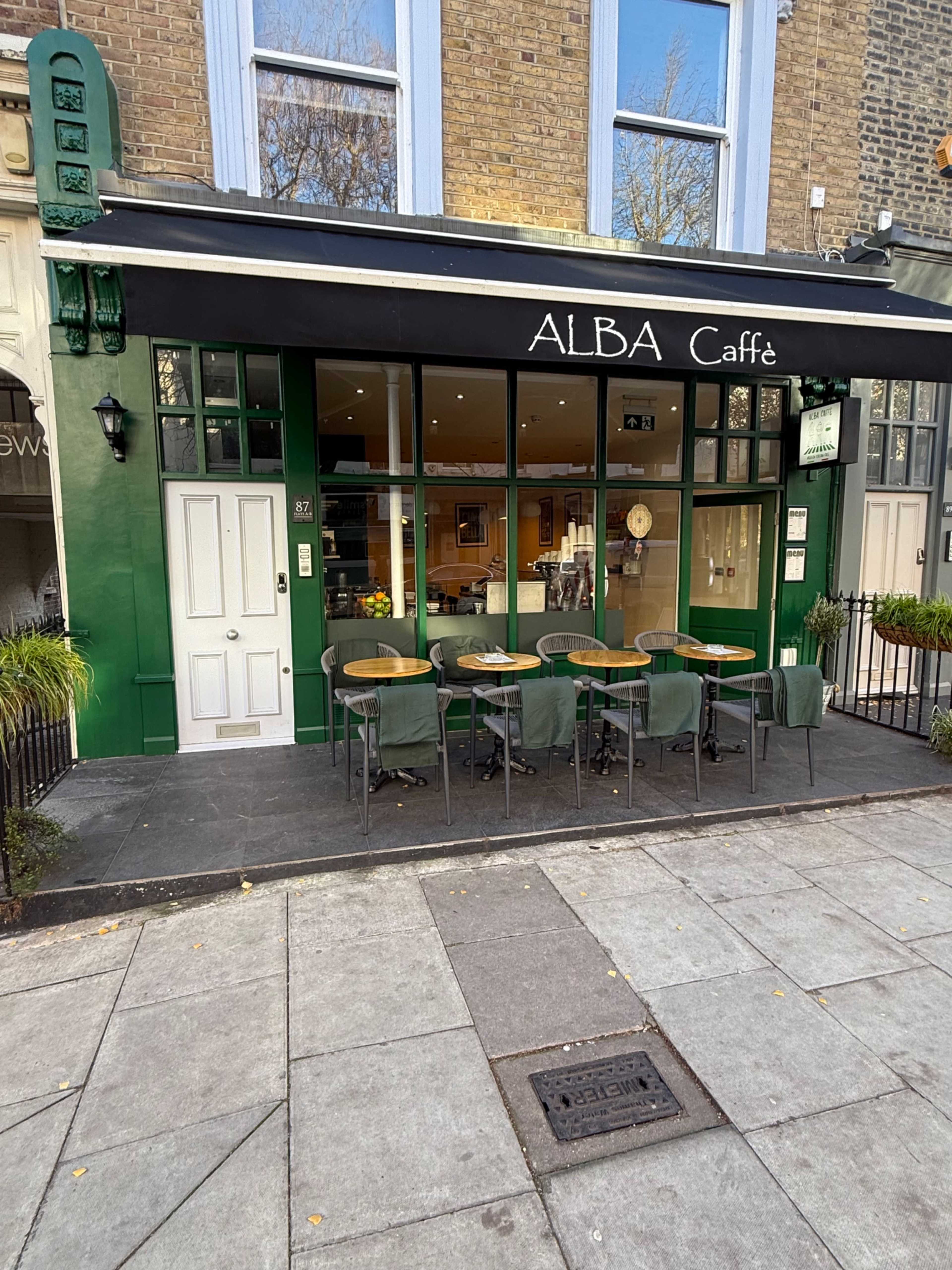 The image shows the exterior of a cafe named "ALBA Caffe," featuring a green facade, large windows, and several outdoor tables with chairs in front.