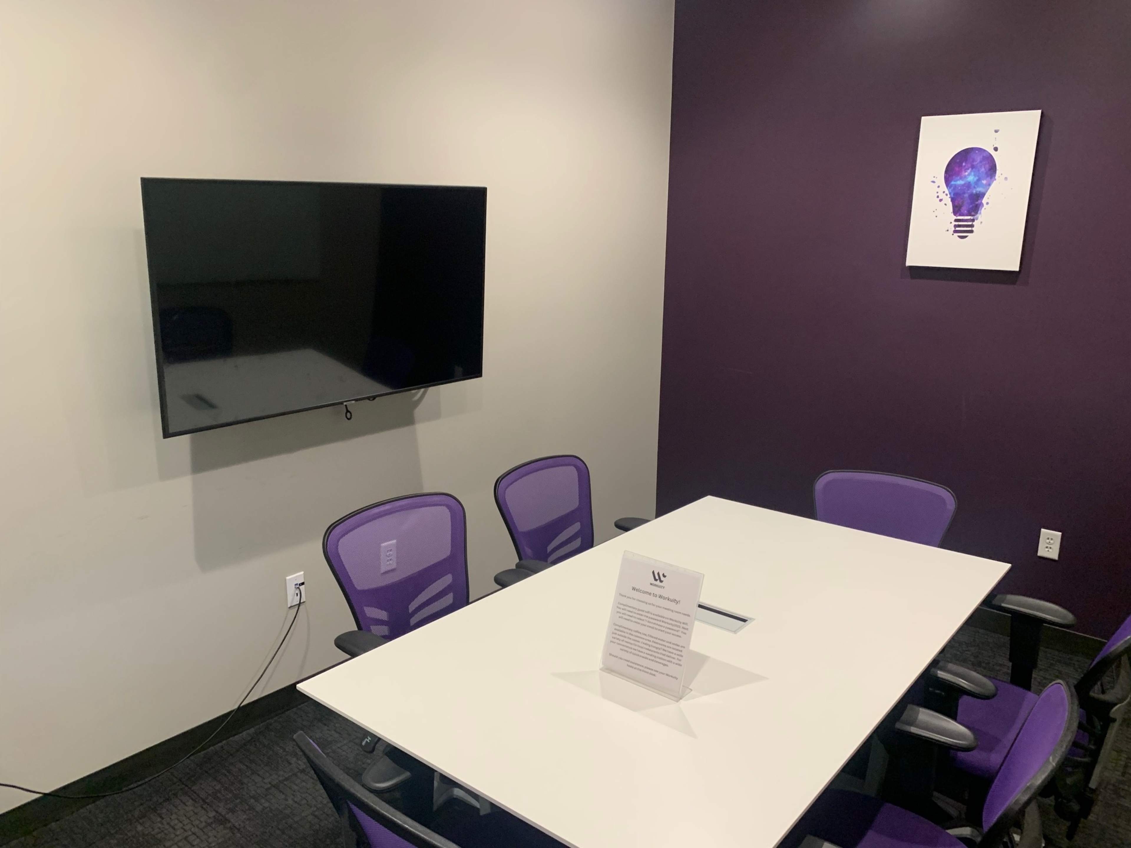 A modern meeting room features a white table surrounded by purple chairs and a wall-mounted television.
