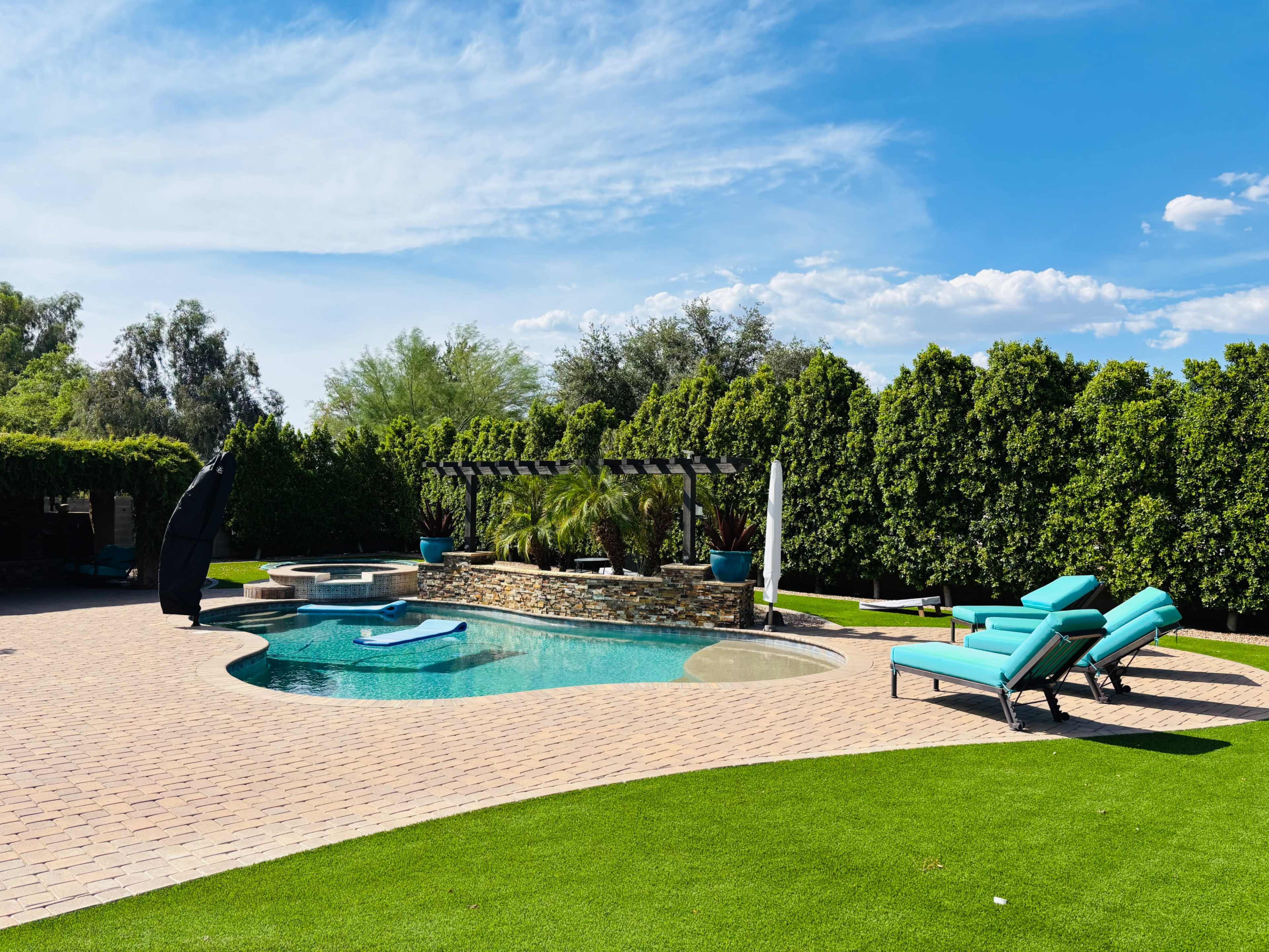 The image shows a residential backyard with a swimming pool, lounge chairs, and well-maintained landscaping under a clear blue sky.
