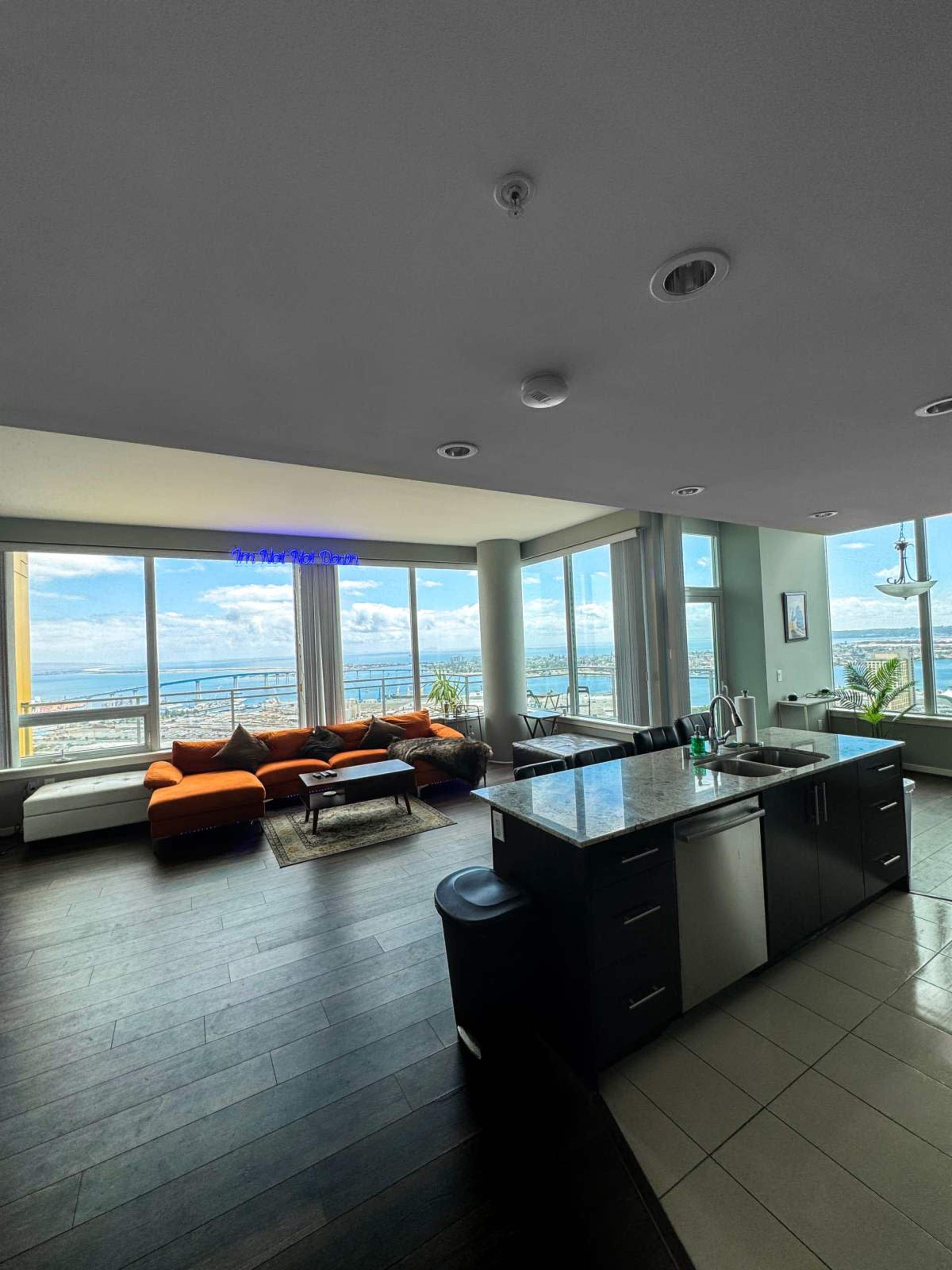 A modern living space with a kitchen area, a large orange sofa, large windows offering a view of the outdoors, and wooden flooring.