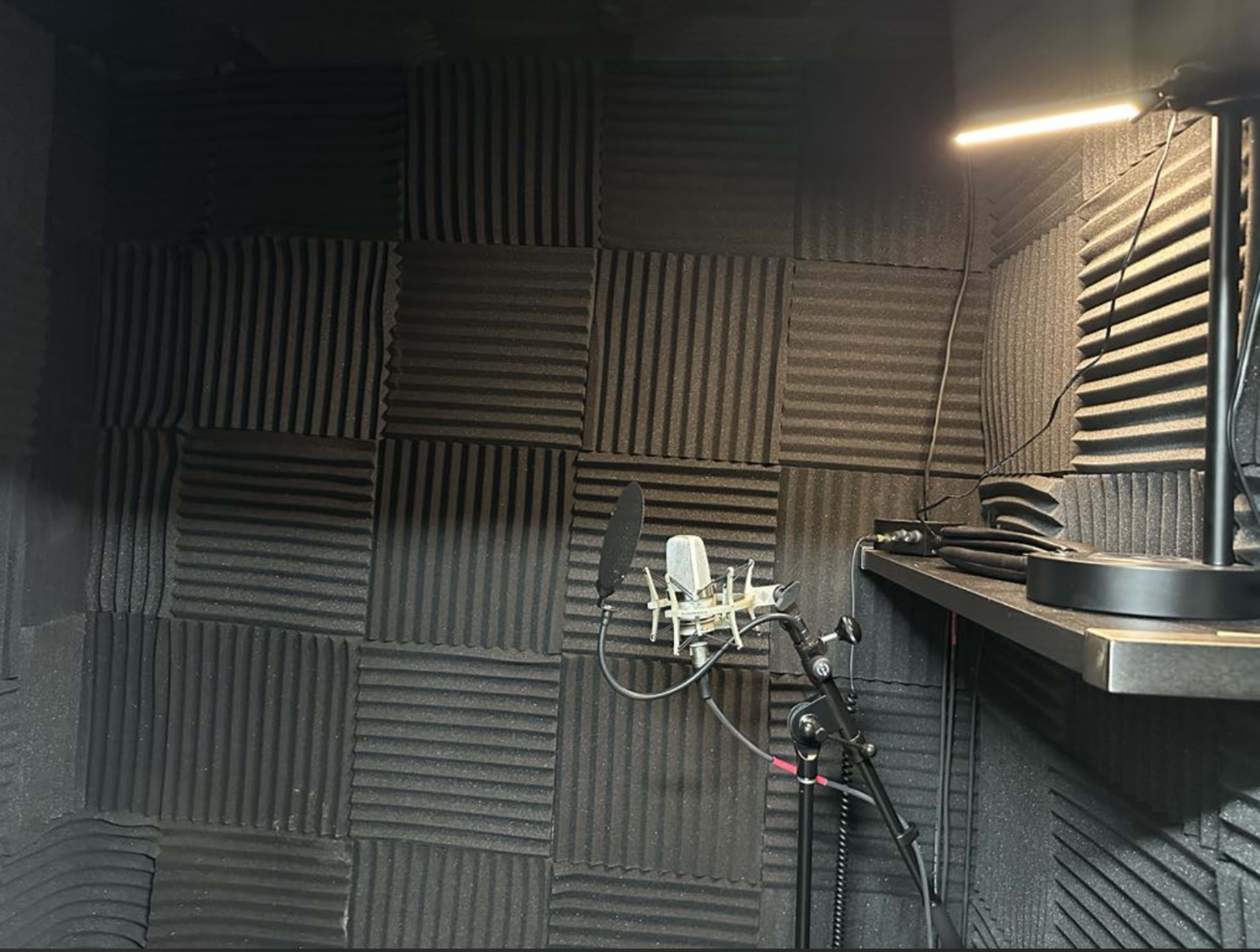 The image shows a soundproof recording booth lined with acoustic foam panels, featuring a microphone set up on a stand and a shelf with equipment.