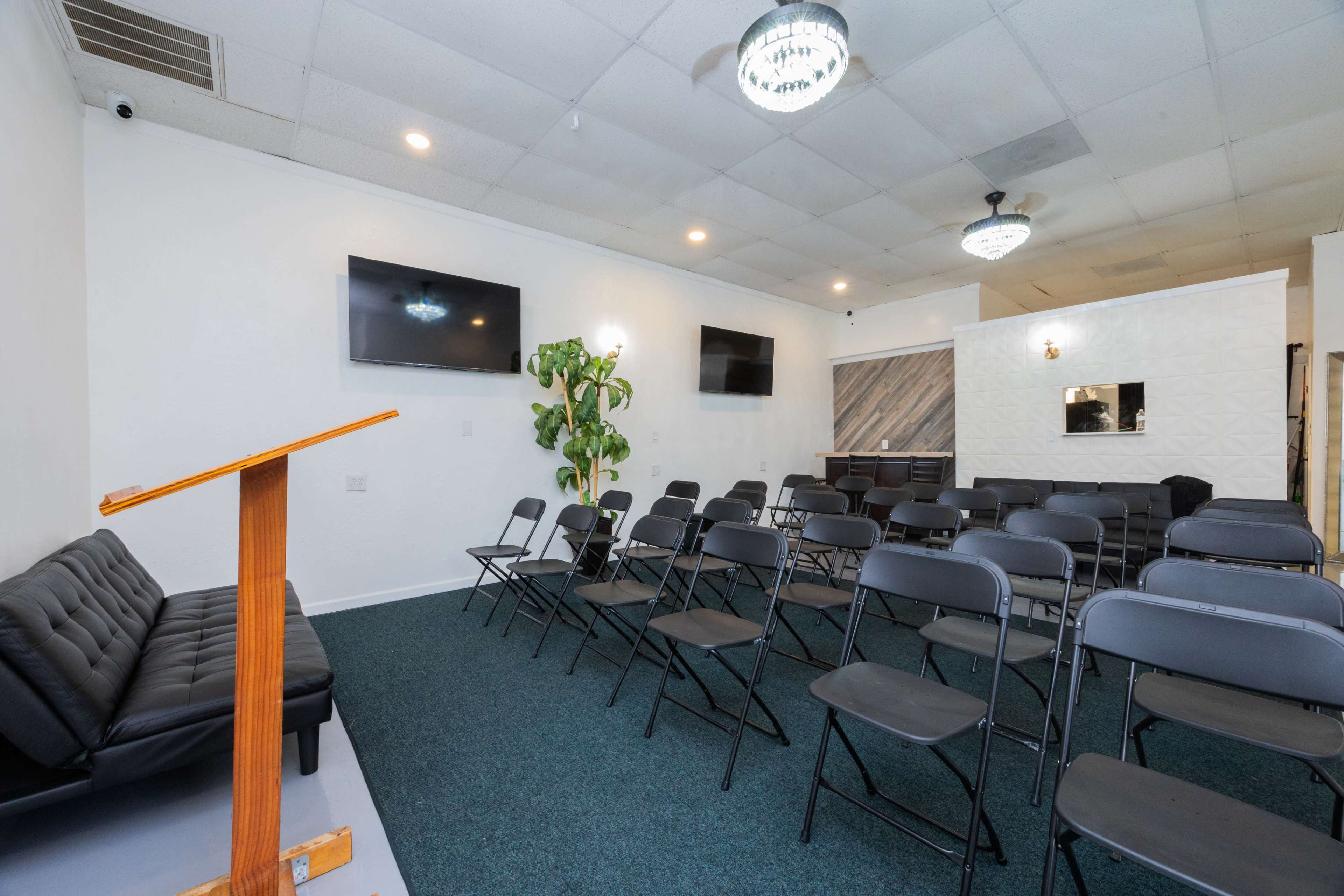 A room set up for a gathering, featuring rows of black folding chairs, a podium, and two wall-mounted TVs.