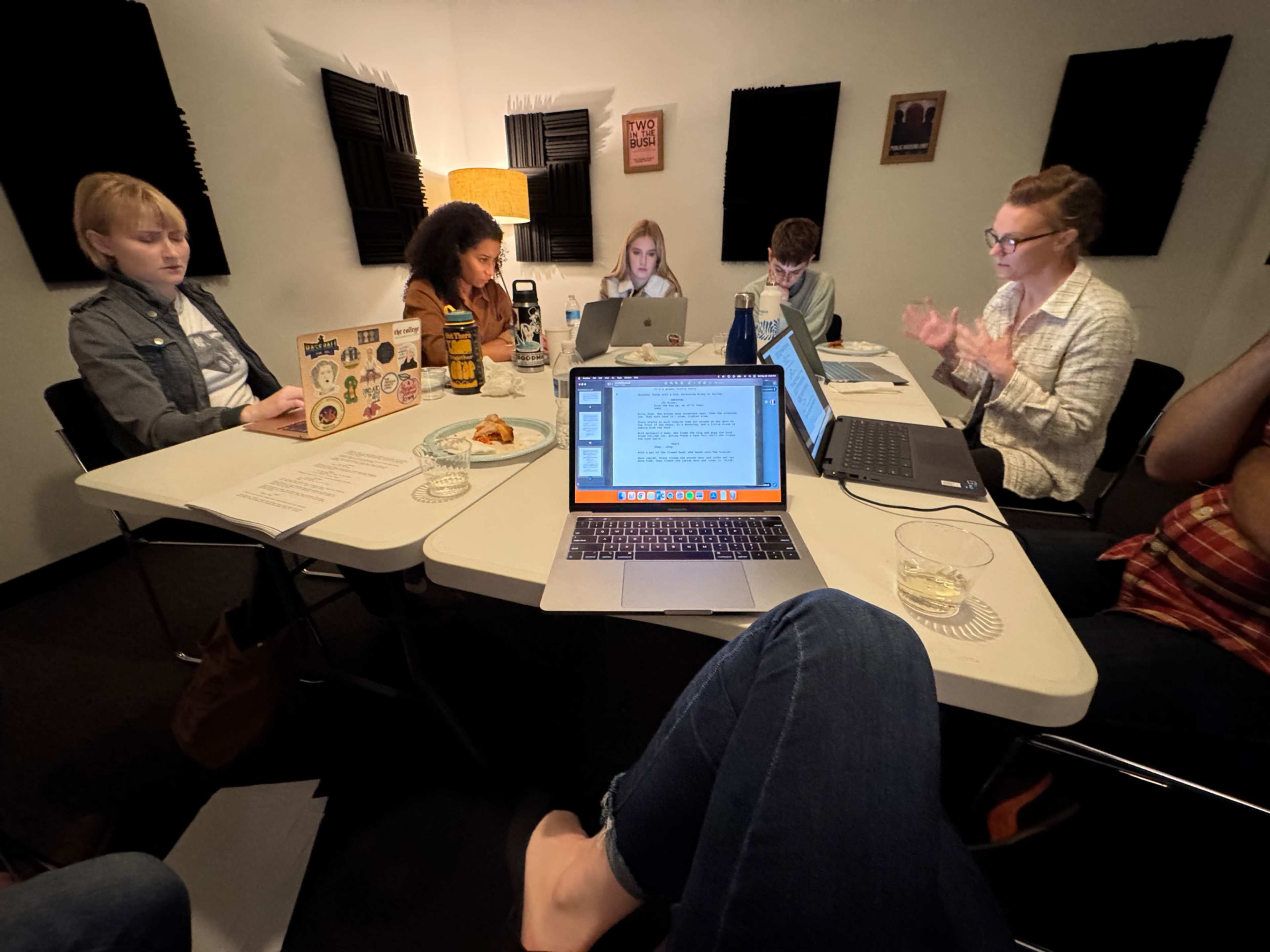 A group of six people is seated around a table in a small room, working on laptops and discussing as they review documents and notes.