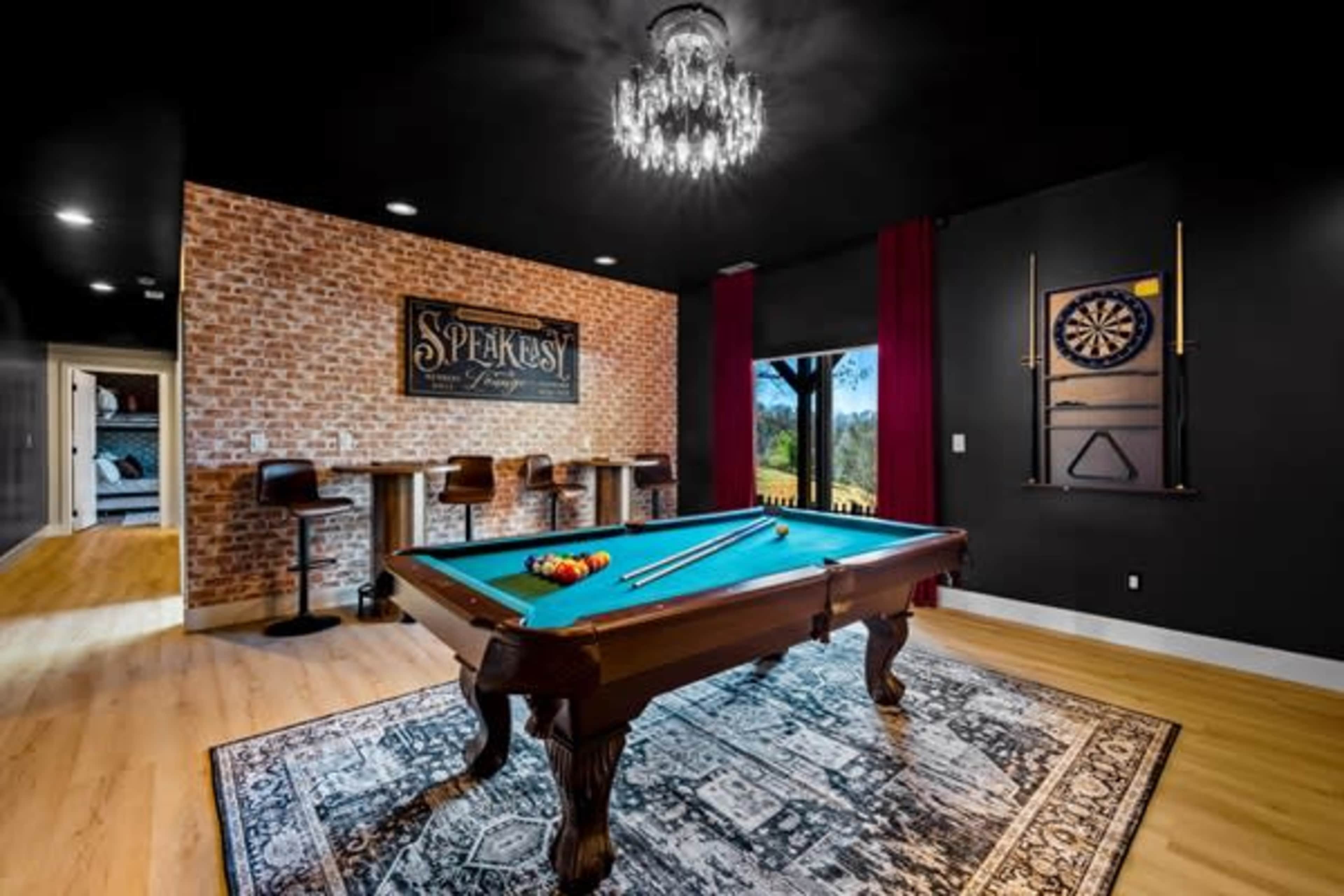 A billiards room features a green pool table, a bar with high stools, and a dartboard against a brick wall, illuminated by a chandelier.
