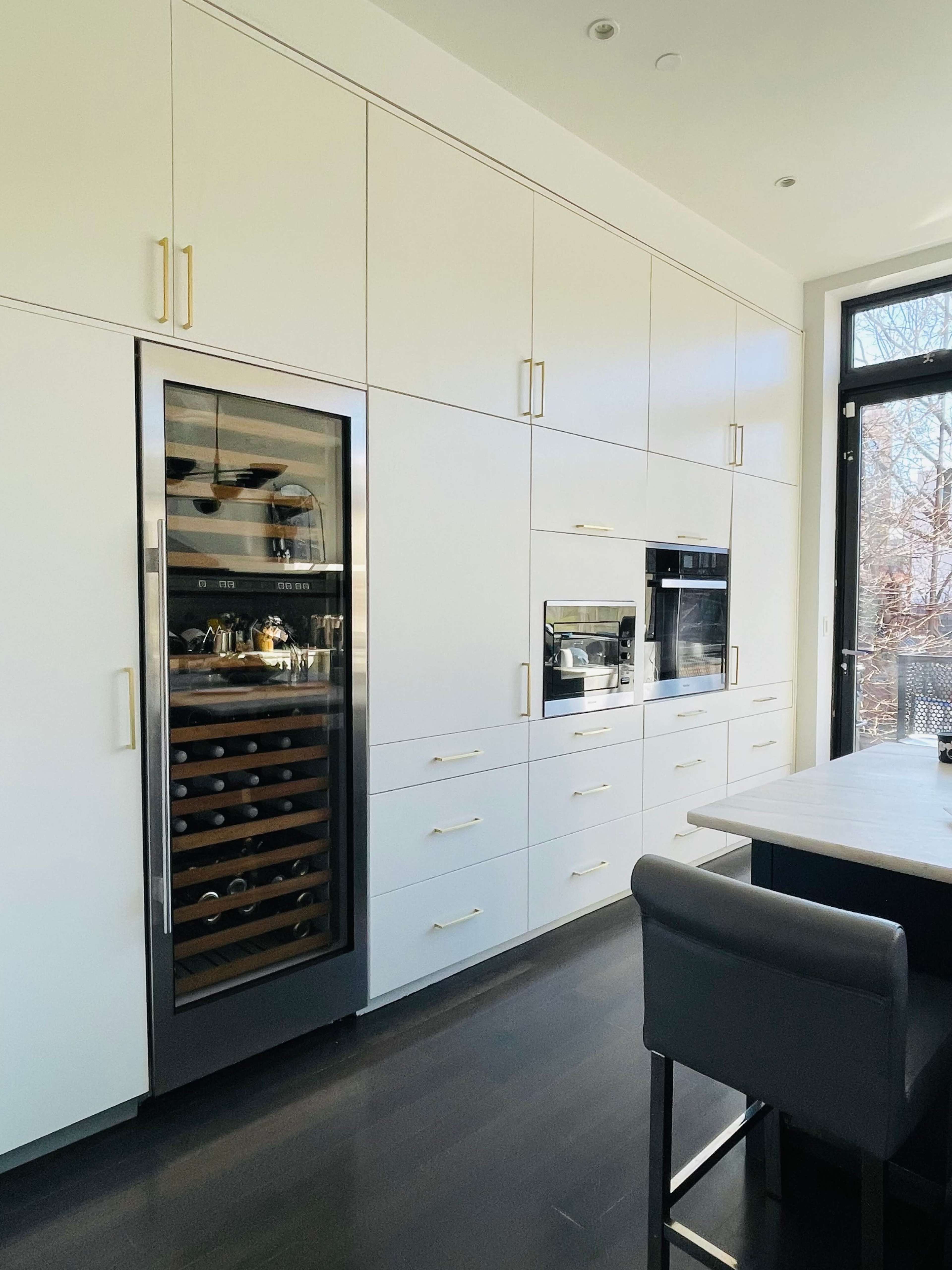 A modern kitchen with white cabinetry, an integrated wine cooler, and built-in appliances, all set against a large window.