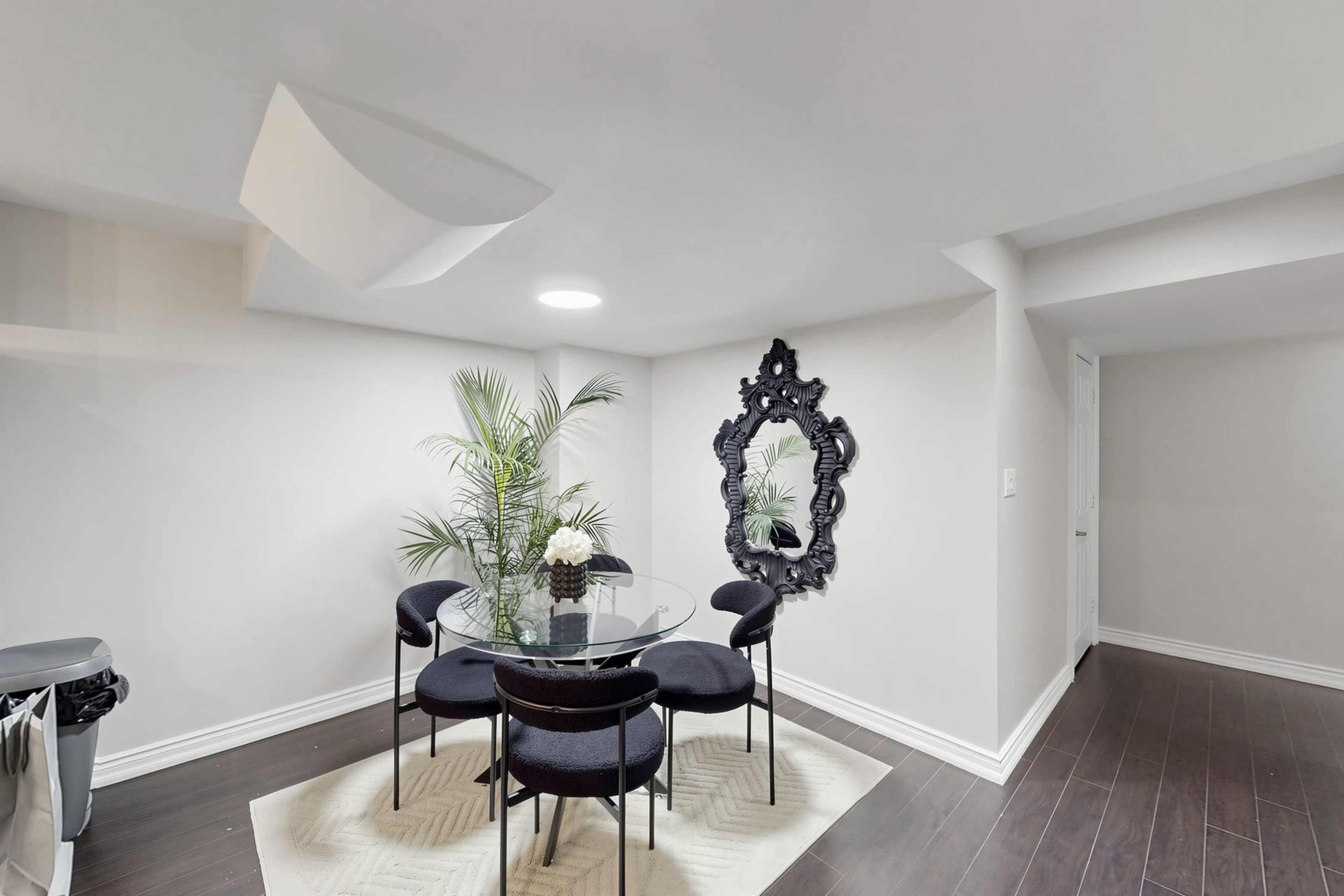 A modern dining area features a glass table surrounded by four black chairs, a decorative mirror on the wall, and a potted plant in the corner.