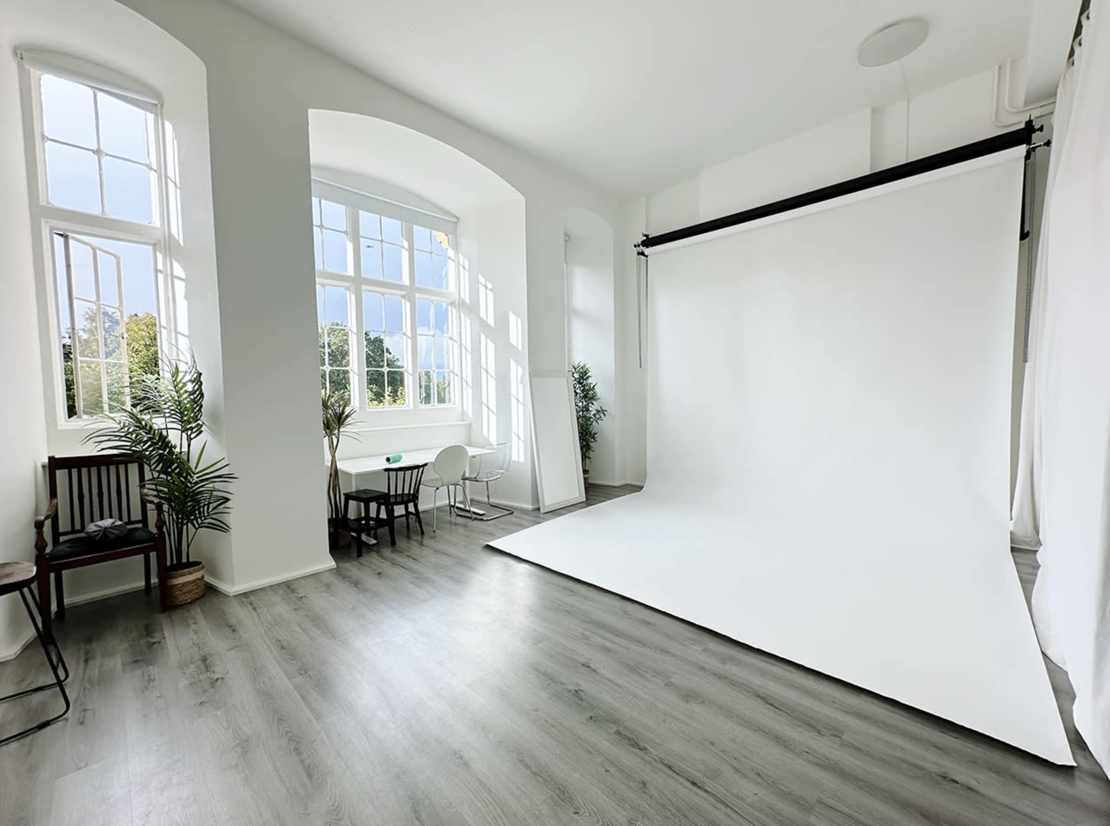 The image shows a bright studio space with large windows, a white backdrop, and minimalist furniture including a chair and a small table.
