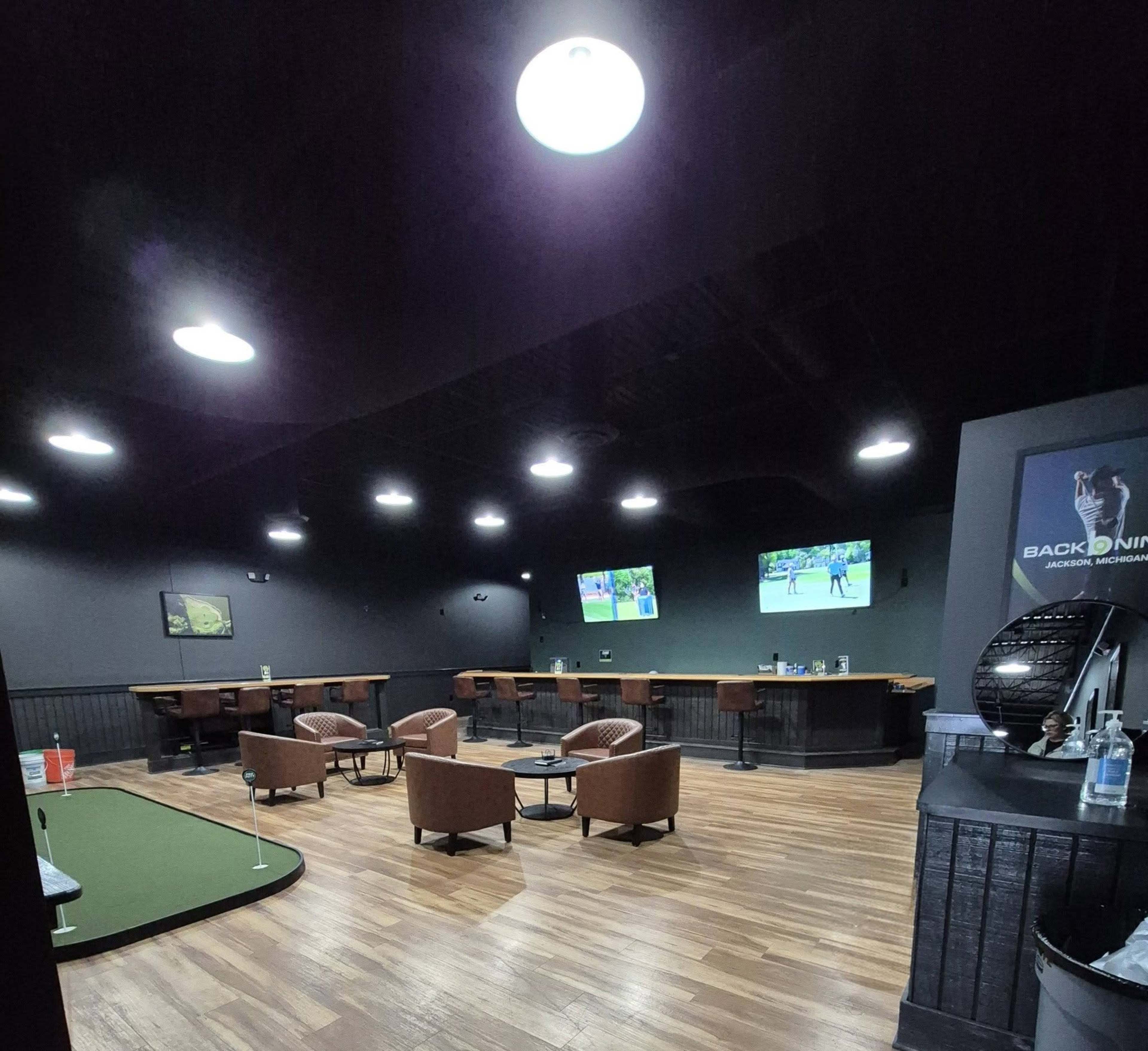 The image shows a darkened lounge area featuring a putting green, two televisions displaying sports, and several brown chairs arranged around a central seating area.
