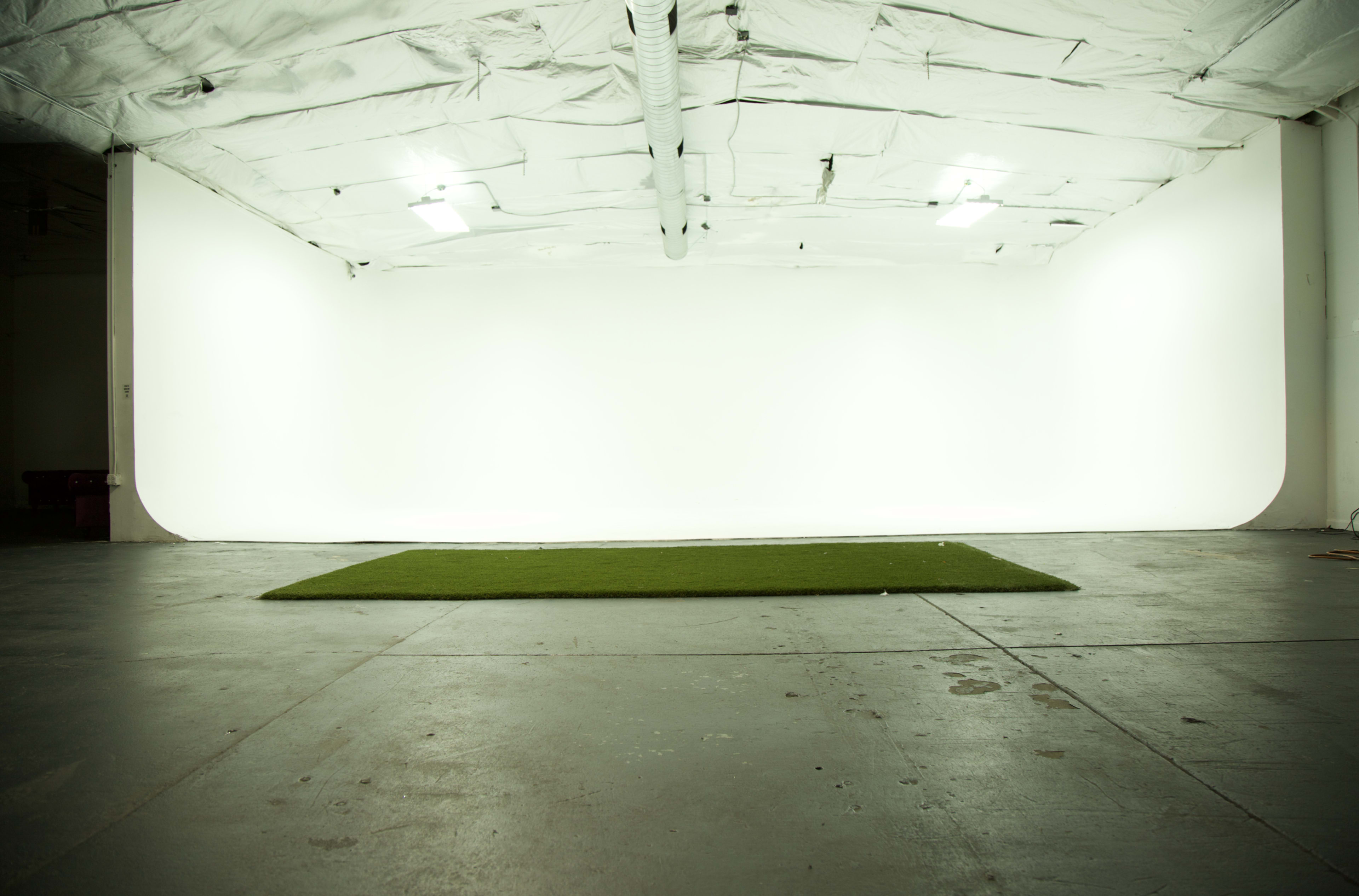 An empty studio space with white walls, a flat green area on the floor, and overhead lighting.