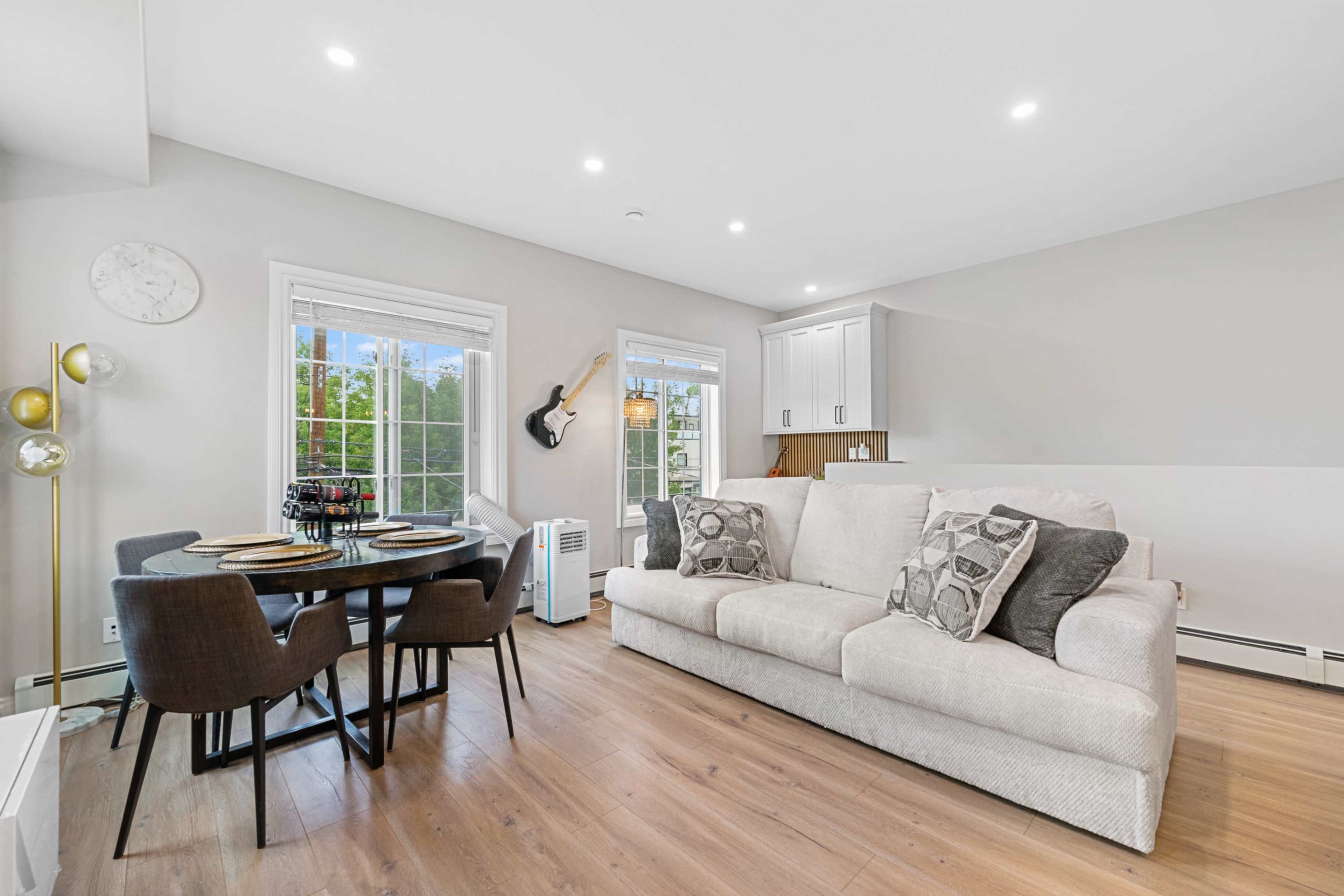 The image shows a modern living area featuring a white sofa, a round dining table with chairs, large windows, and a guitar mounted on the wall.