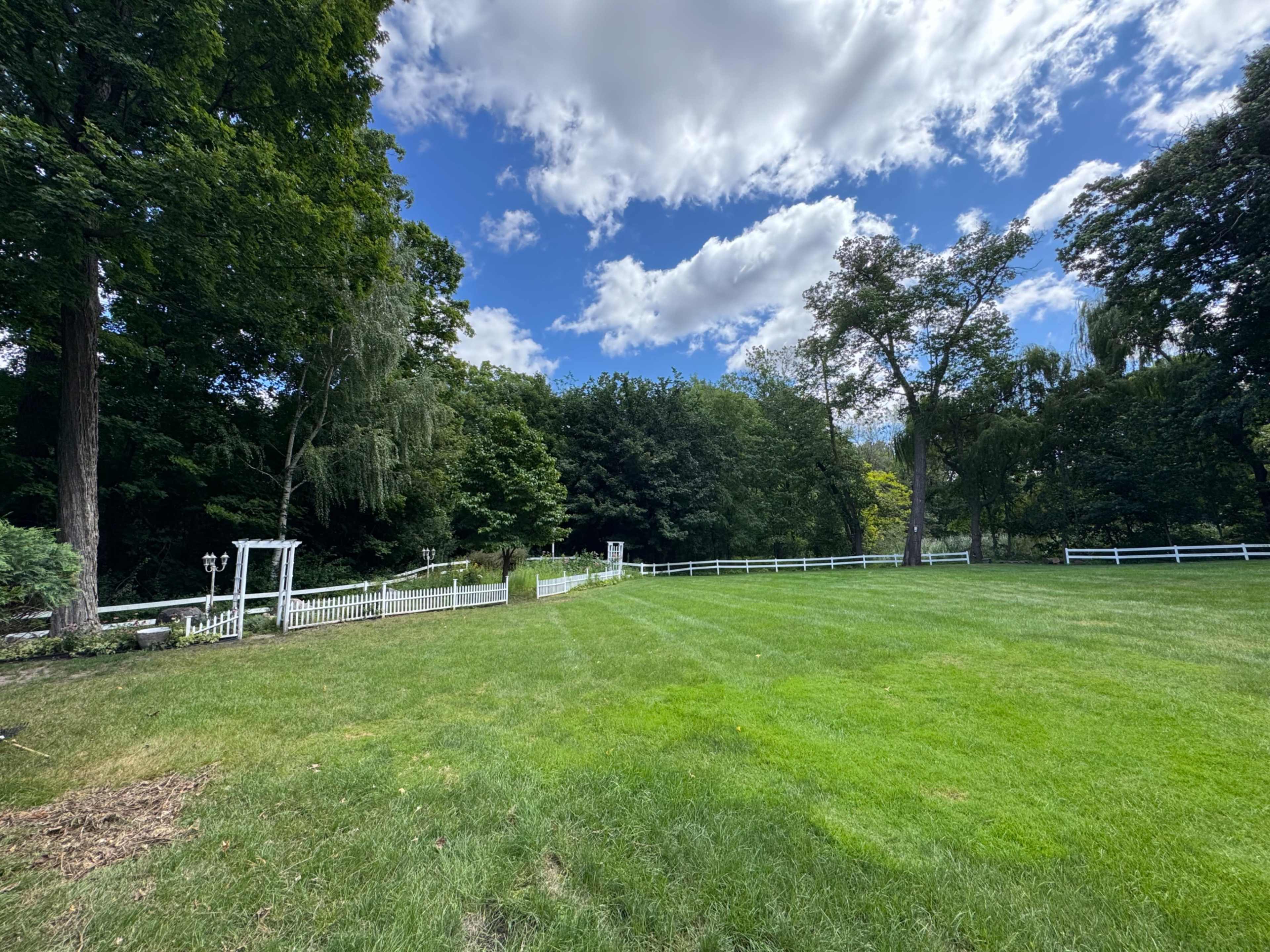 The image shows a green, open lawn bordered by trees with a white fence and a gazebo in the background under a partly cloudy sky.