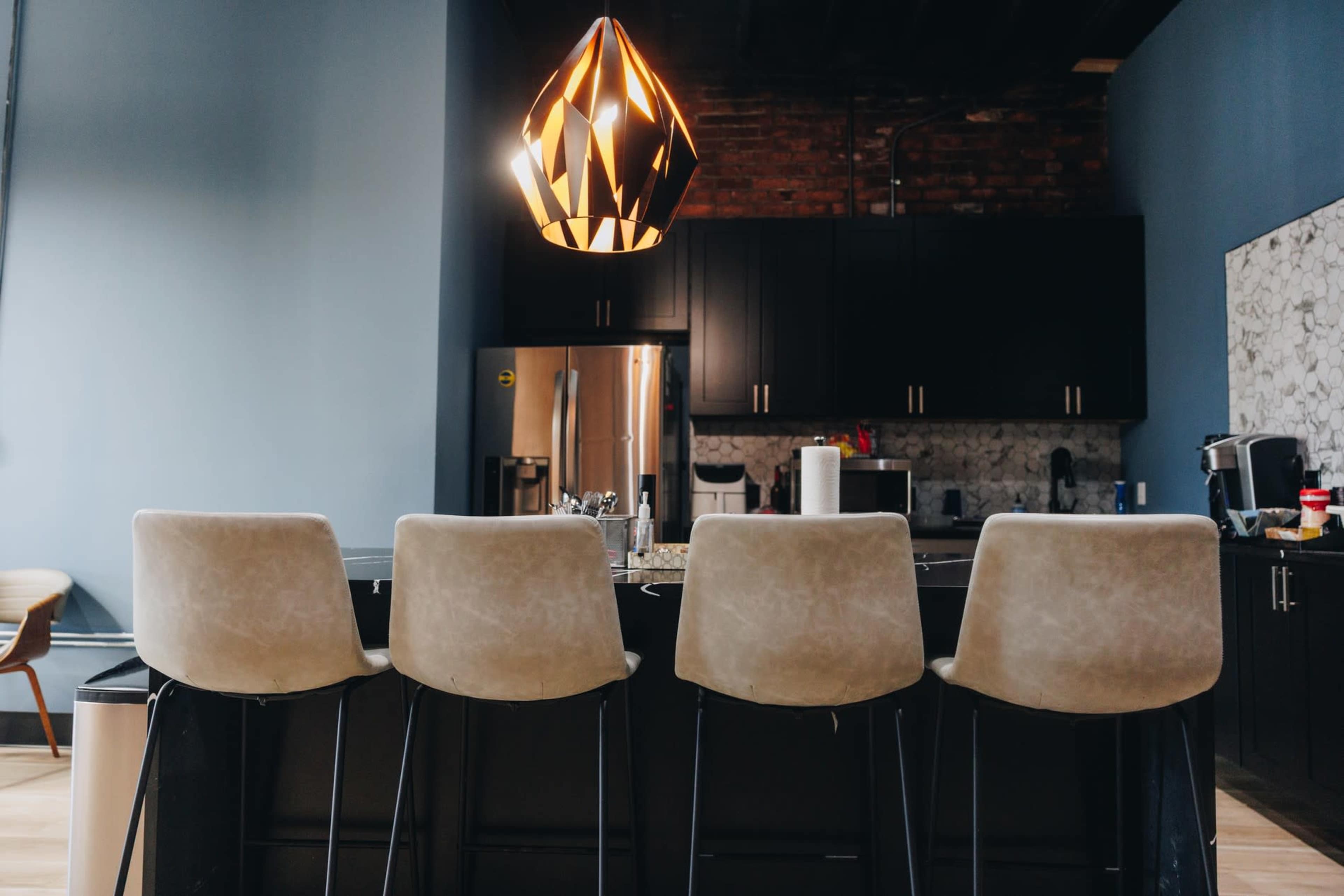 A minimalist kitchen features a bar with four beige stools and a contemporary pendant light hanging above.