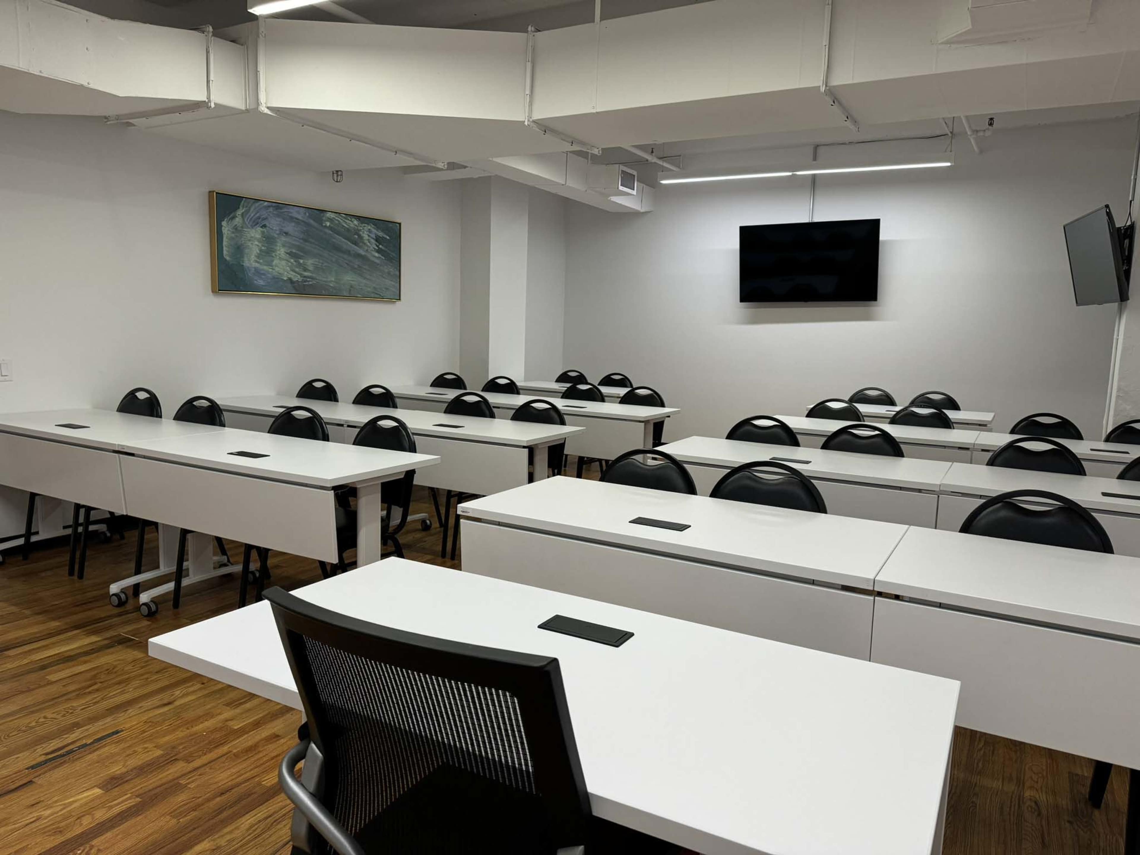 A modern classroom with multiple rows of white lecture tables and black chairs, equipped with a television screen and wall art.