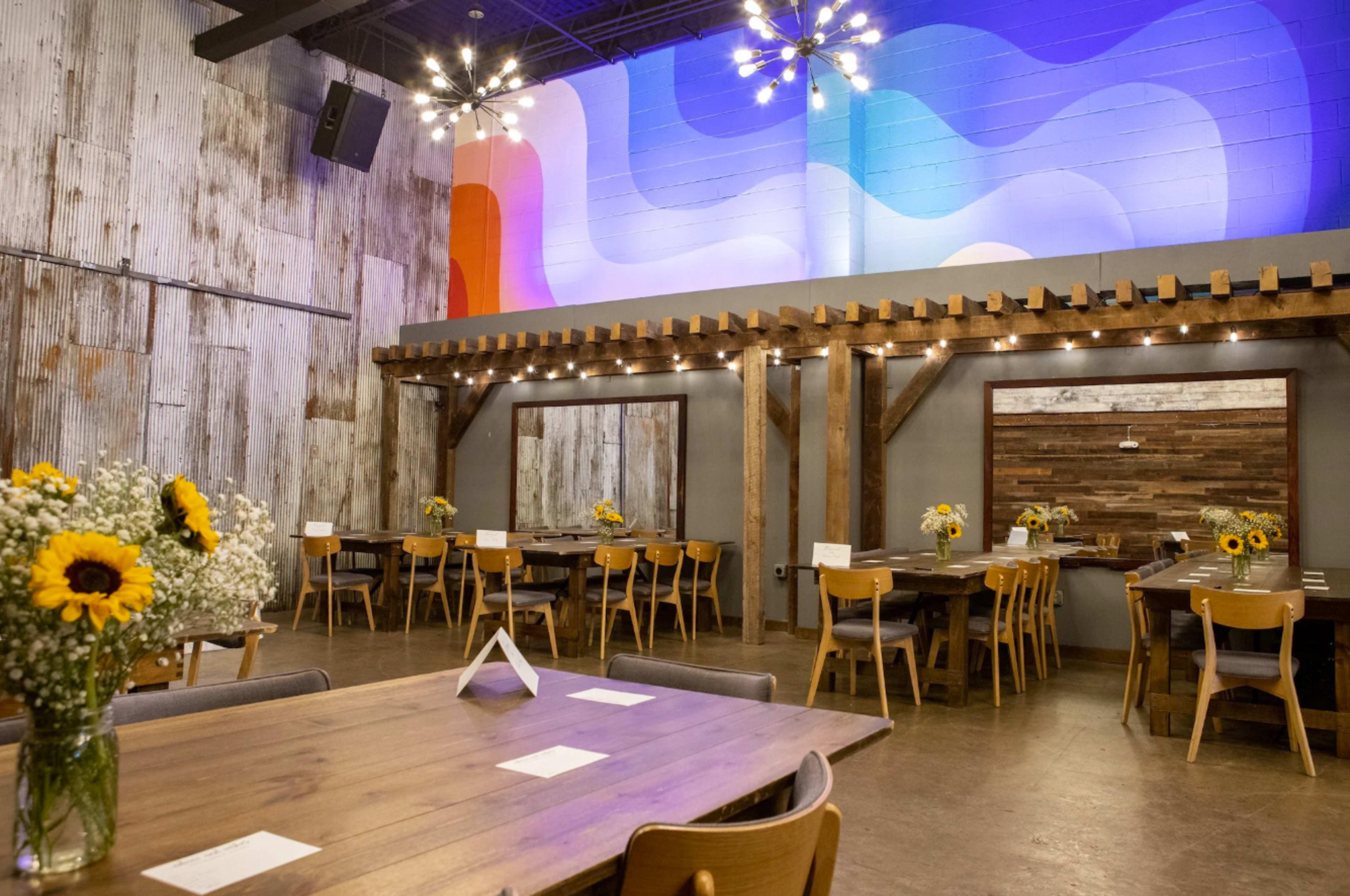 The image shows a spacious dining area with wooden tables, chairs, and floral centerpieces, featuring a colorful abstract mural on the wall.
