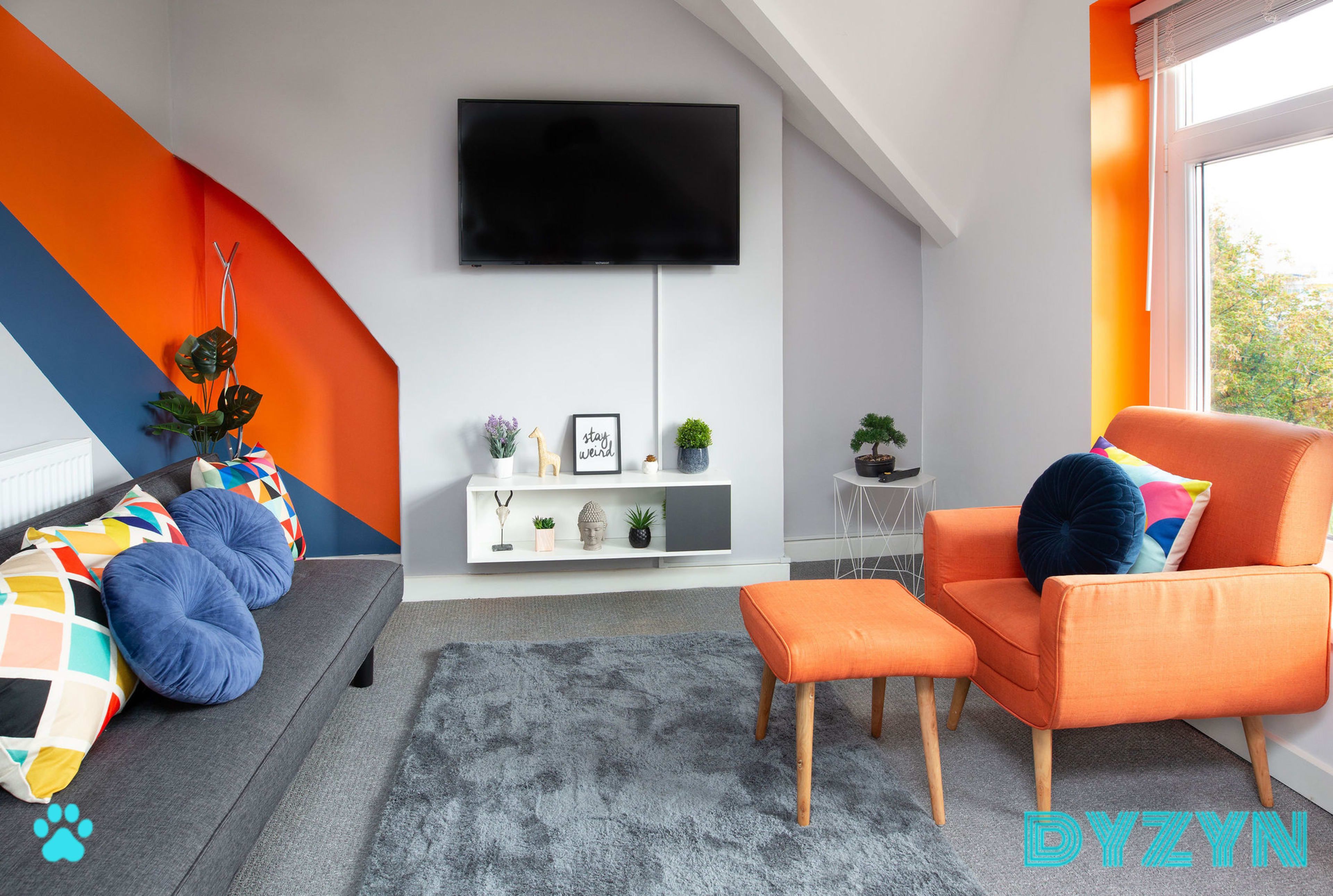 The scene features a modern living room with a gray sofa, an orange accent chair, a white shelf mounted on the wall holding decorative items, and a large flat-screen TV.