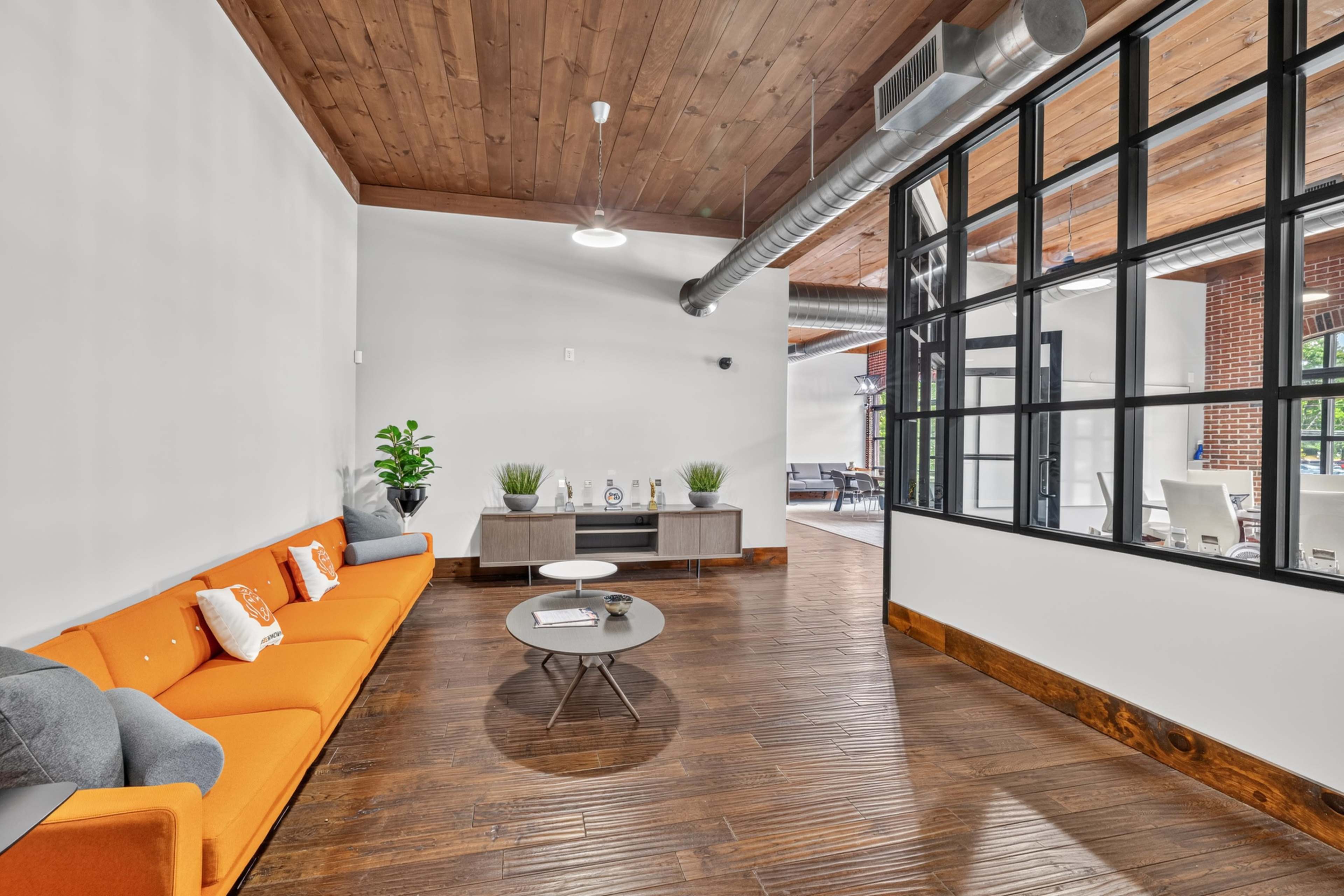 The image shows a modern interior space featuring a bright orange sofa, a coffee table, and large windows overlooking an outdoor area.
