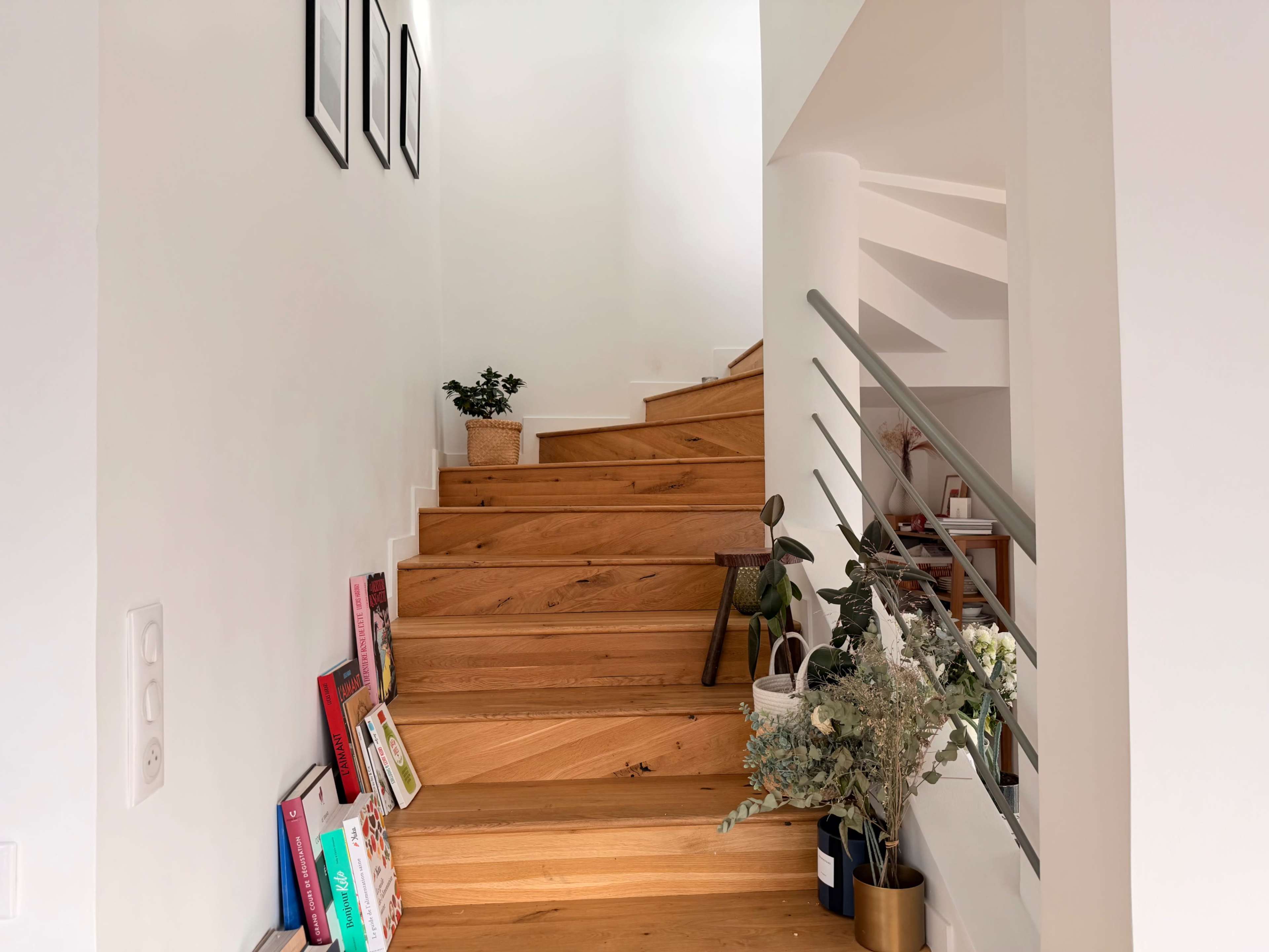 The image shows a well-lit staircase with wooden steps, surrounded by a white wall and framed pictures, along with plants and books arranged on the side.