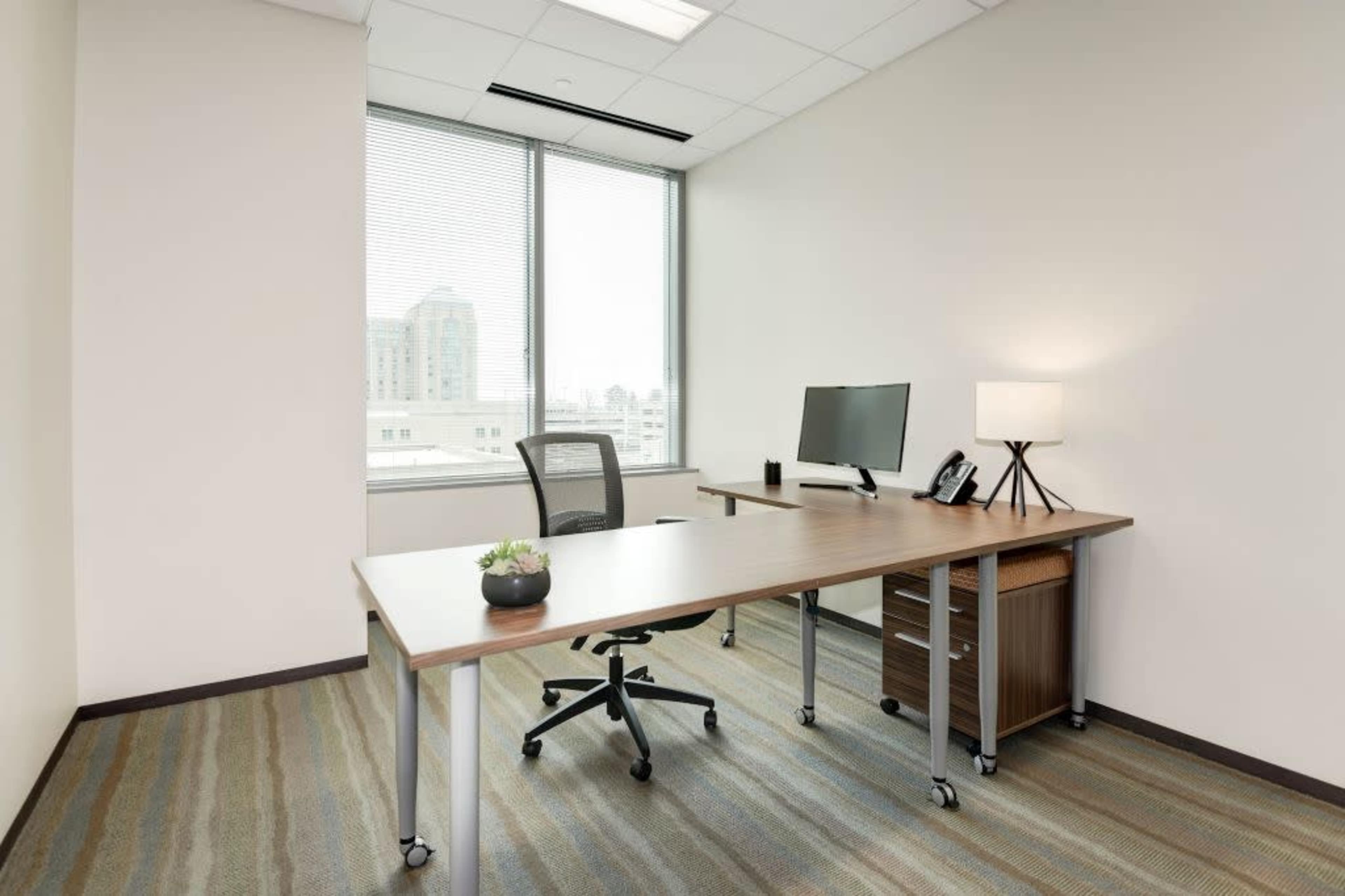 A modern office space featuring a wooden desk, a computer monitor, a small lamp, and an office chair near a large window with city views.