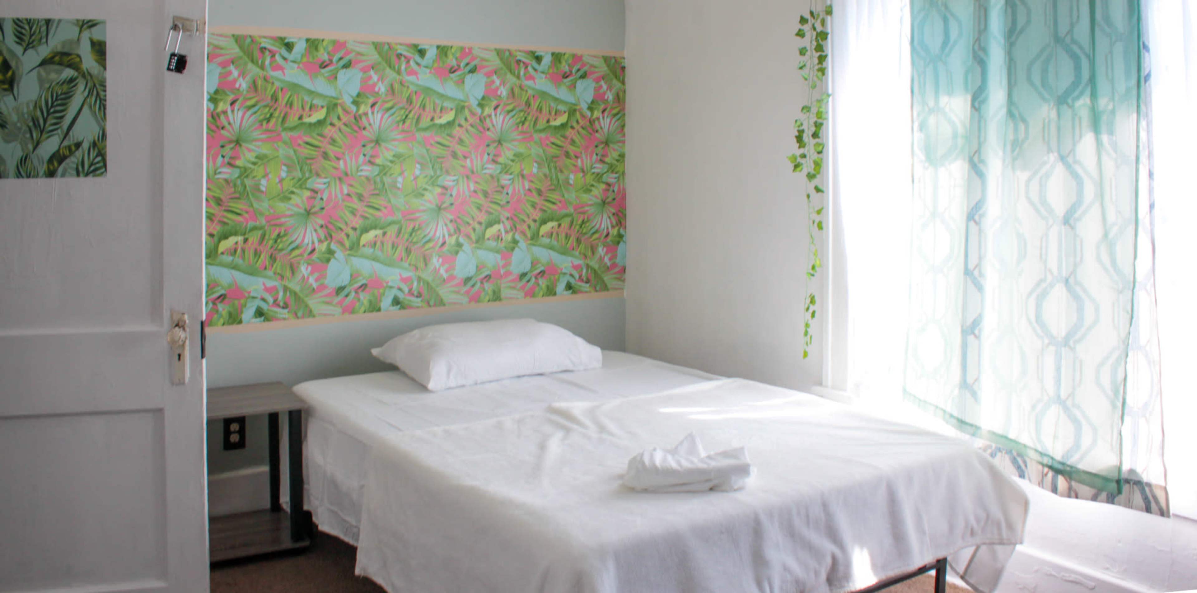 A simple bedroom with a white bed, a decorative wall featuring tropical patterns, and a window covered with sheer curtains.