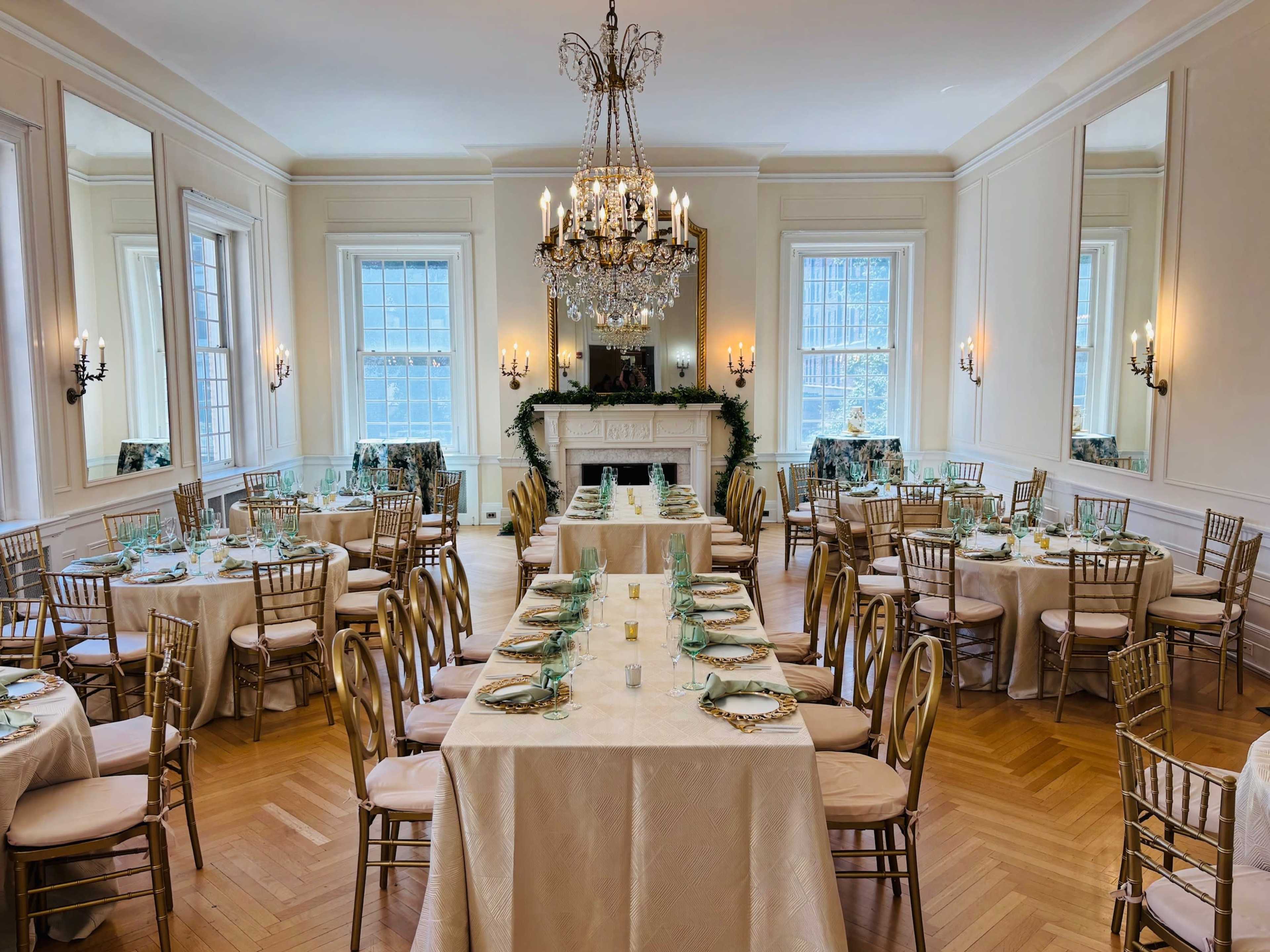 The elegant dining room features several tables set with place settings, surrounded by gold Chiavari chairs and adorned with a chandelier overhead.
