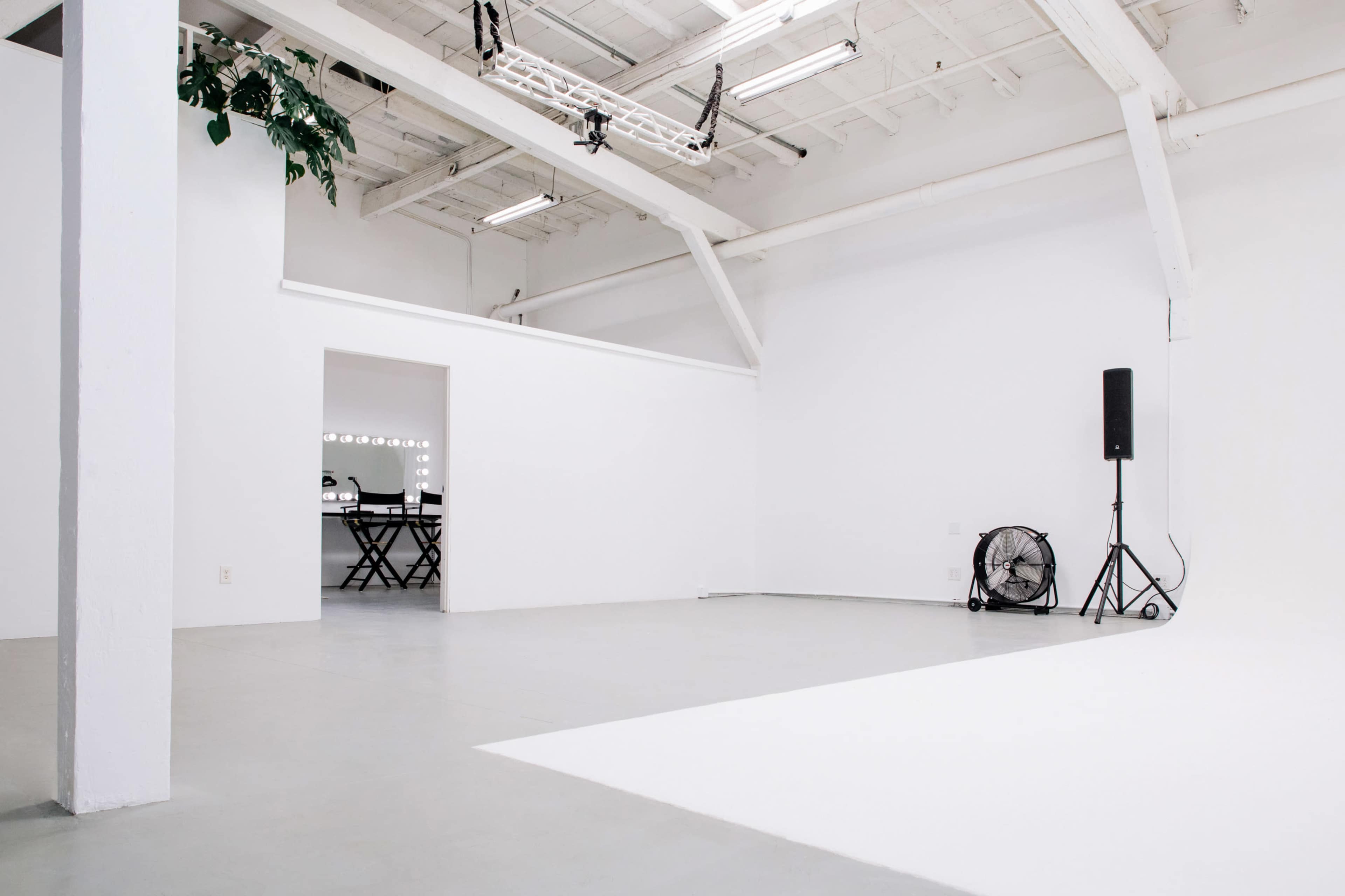 A spacious, white room with a large open area, a wall-mounted speaker, a fan, and a doorway leading to a separate space with a table and chairs.