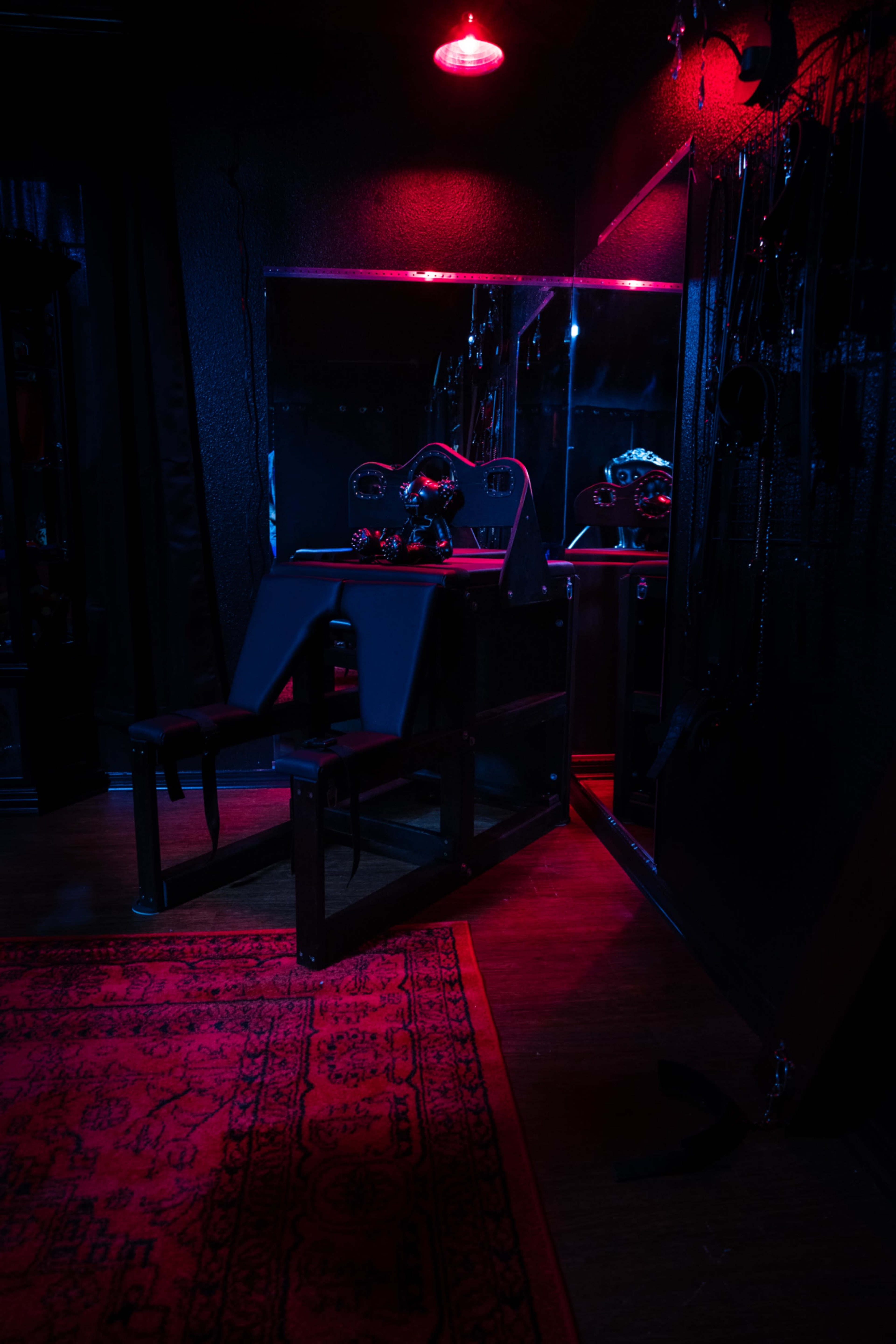 The image shows a dimly lit room with a red glowing light, featuring a black adjustable chair, a mirror, and various items hanging on the walls.