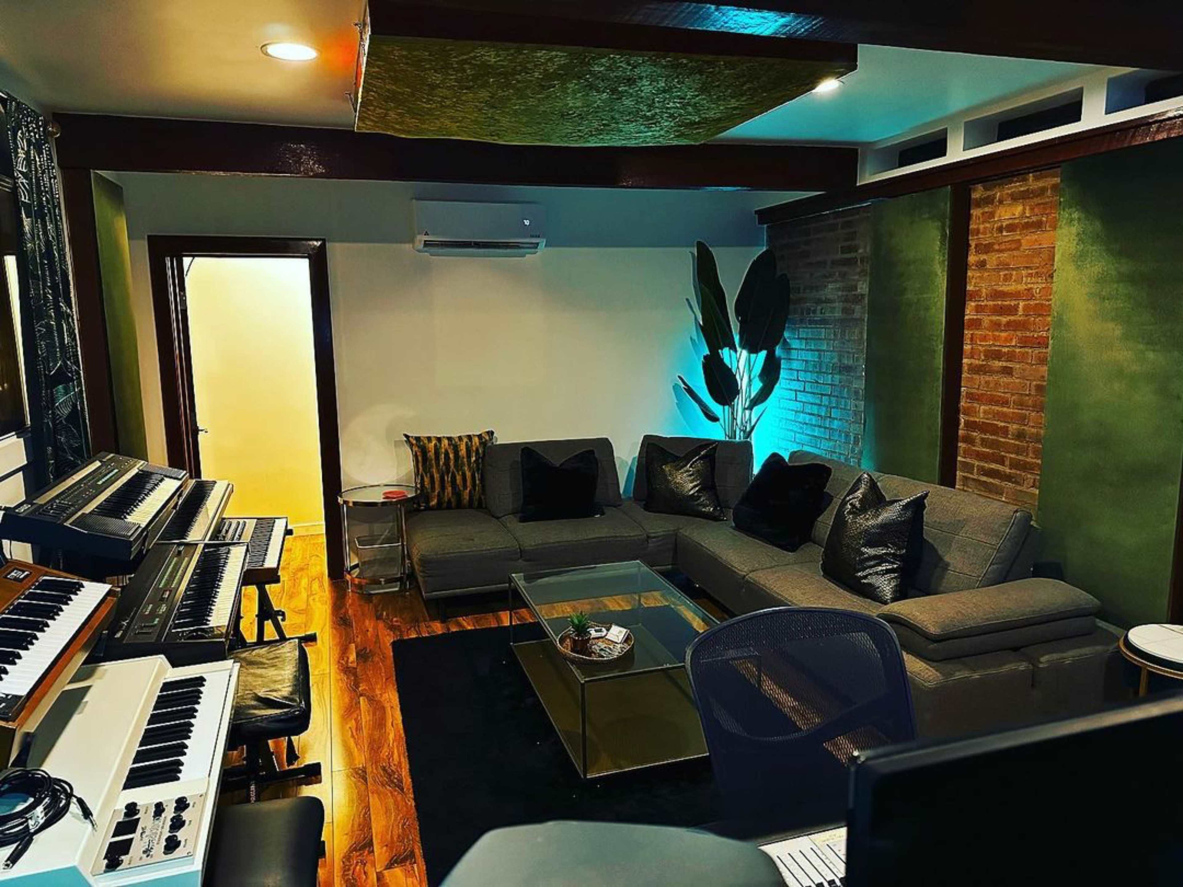 The image shows a music studio living space featuring a sectional sofa, a glass coffee table, and a keyboard setup against a brick wall.