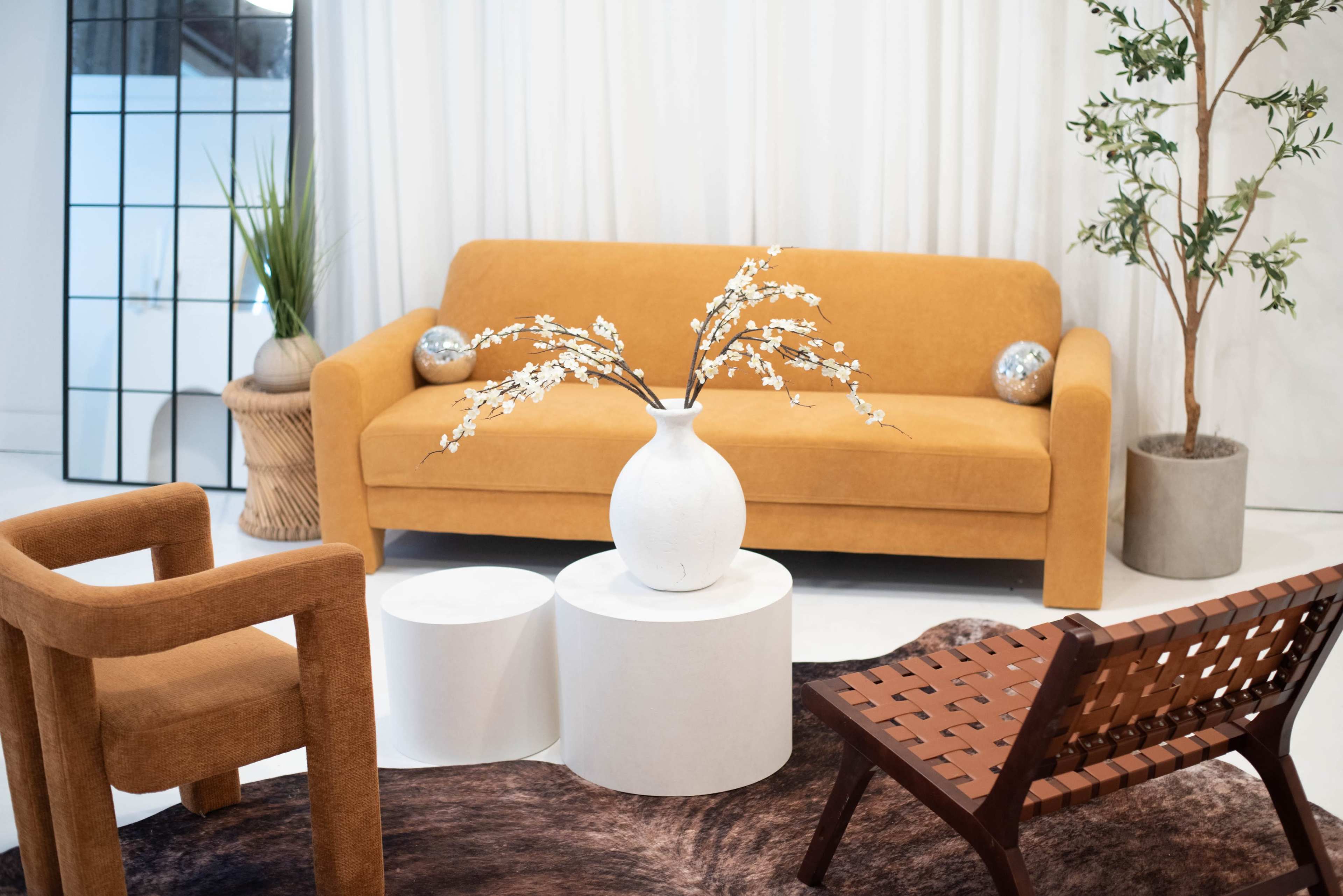 The scene features a cozy living space that includes an orange sofa, a white round vase with decorative branches, and various seating options arranged around a patterned rug.