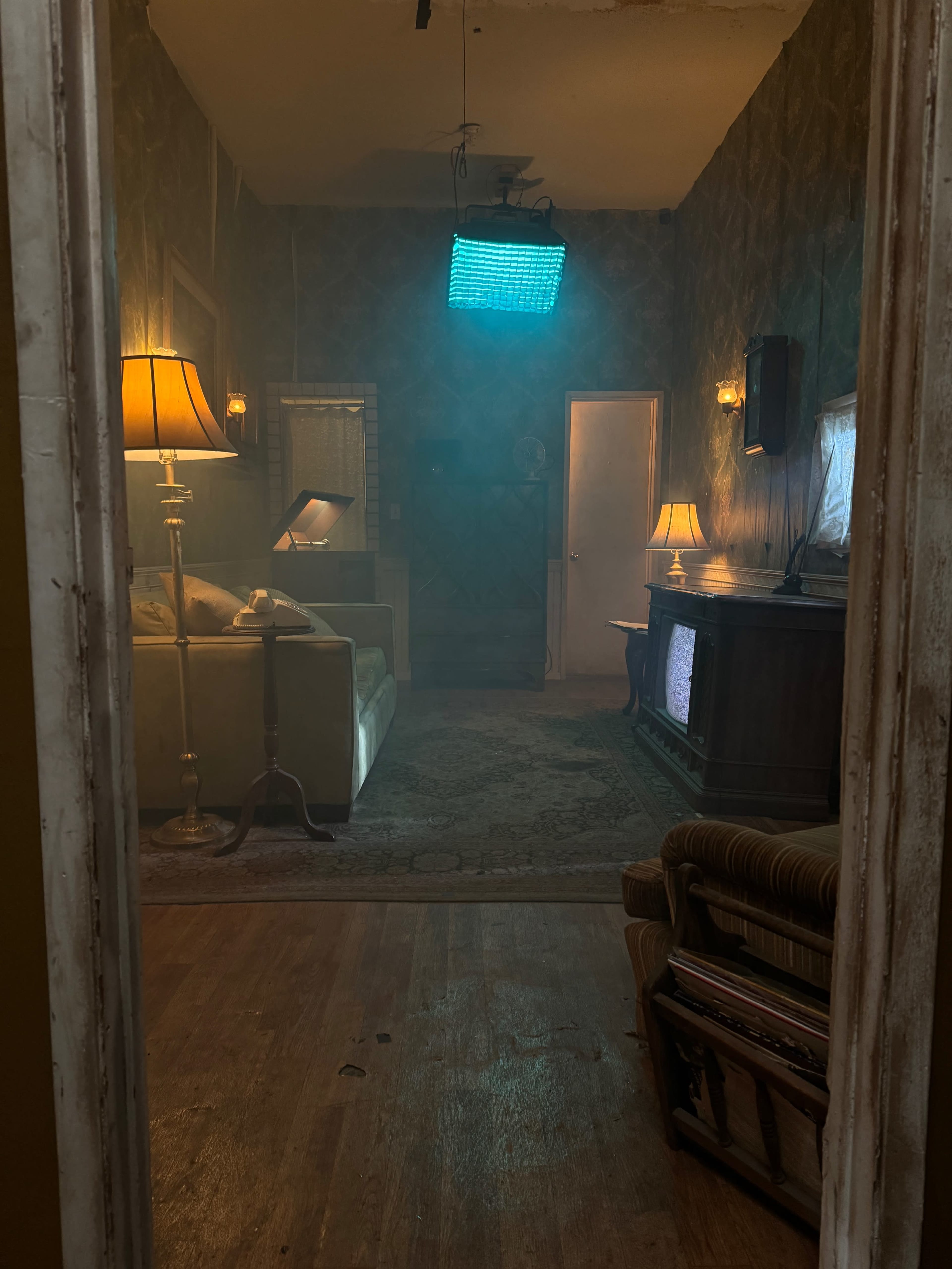 A dimly lit living room features a sofa, side tables with lamps, a vintage television, and a blue light source hanging from the ceiling.
