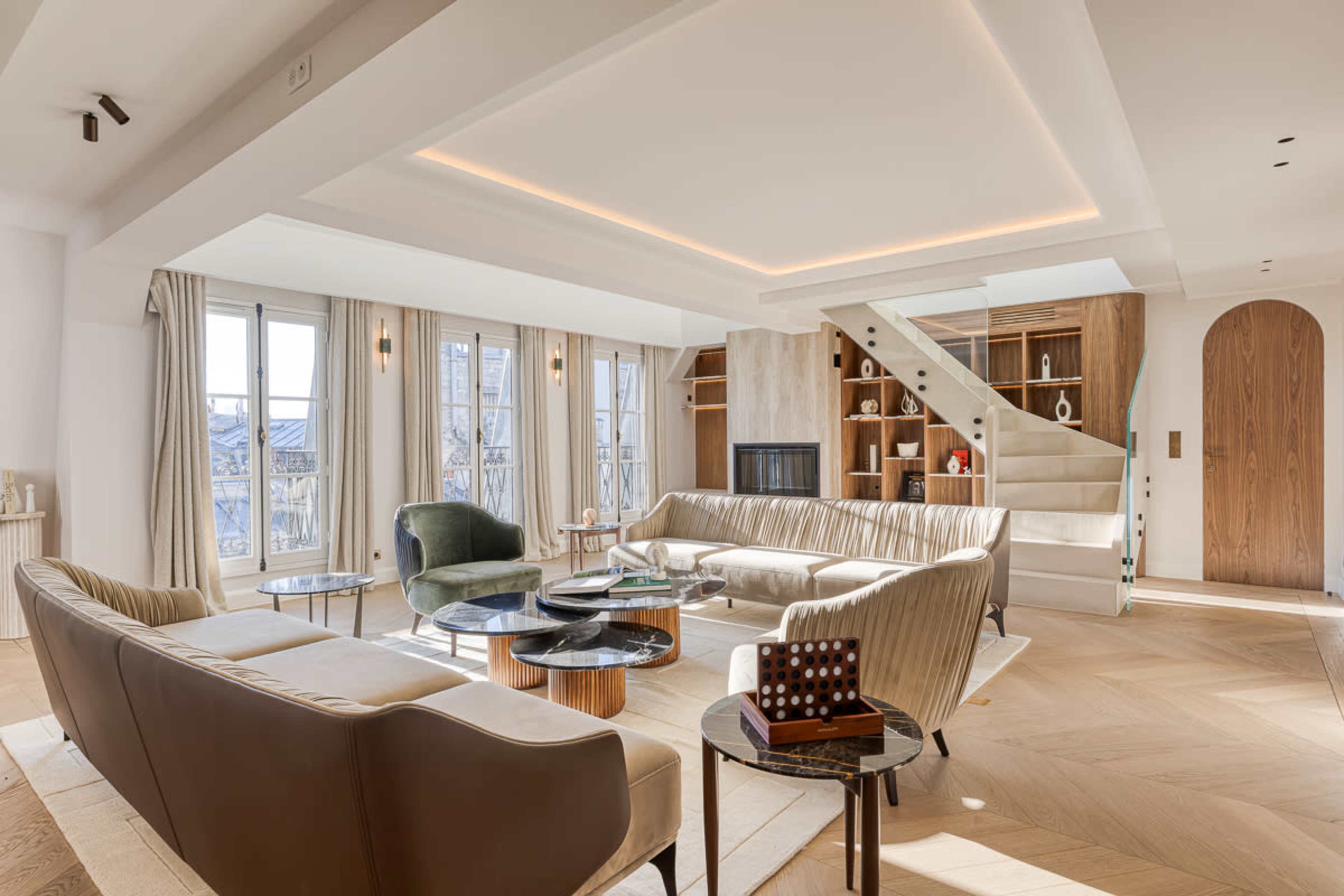 The image shows a modern living room with large windows, featuring a mix of seating options, a staircase, and a wooden shelving unit.