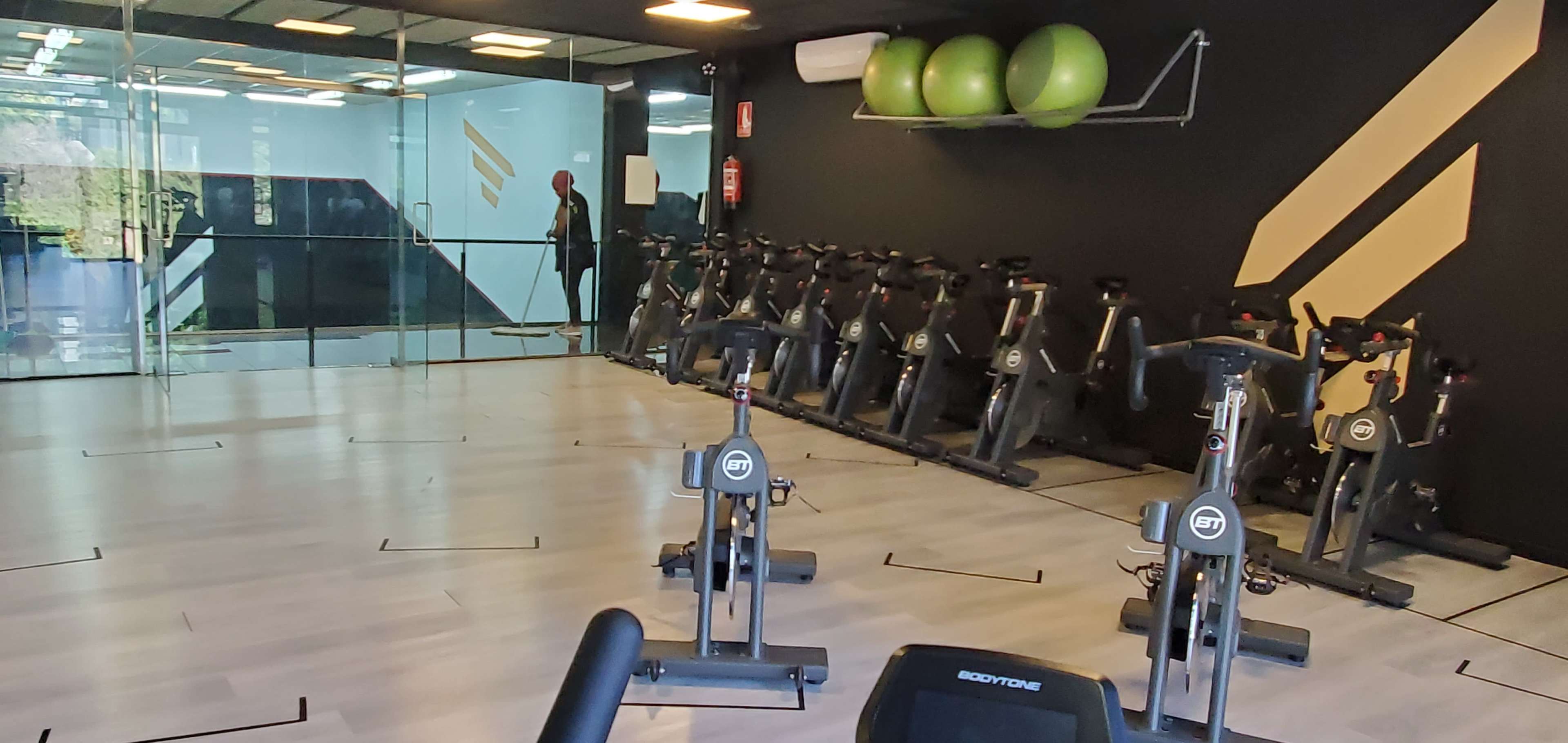 A modern gym studio with rows of stationary bikes and a glass wall separating it from another area, accented by exercise balls on the wall.