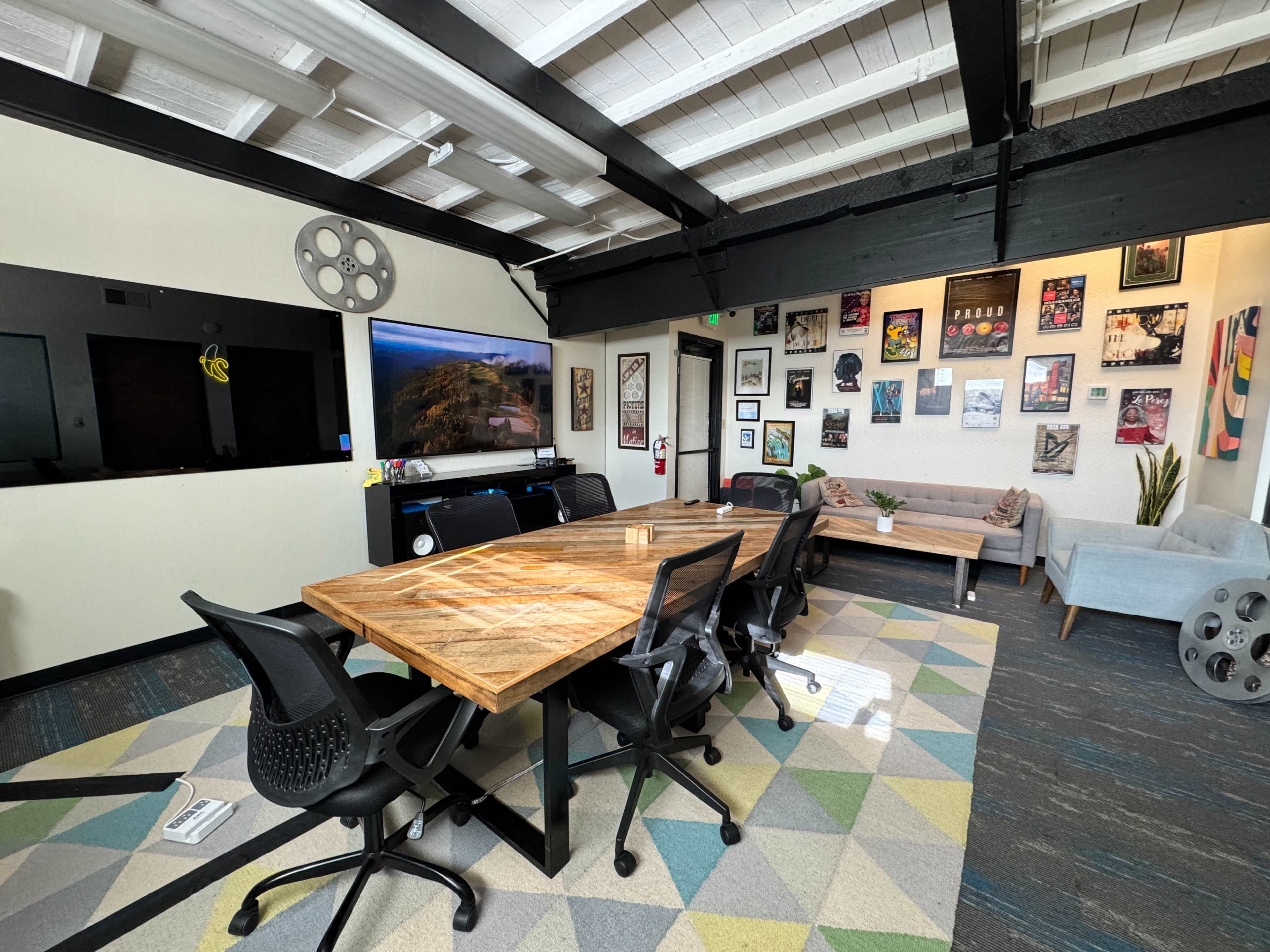 The image shows a modern meeting room with a large wooden table surrounded by black chairs, a television on the wall, and various framed artworks displayed around the room.