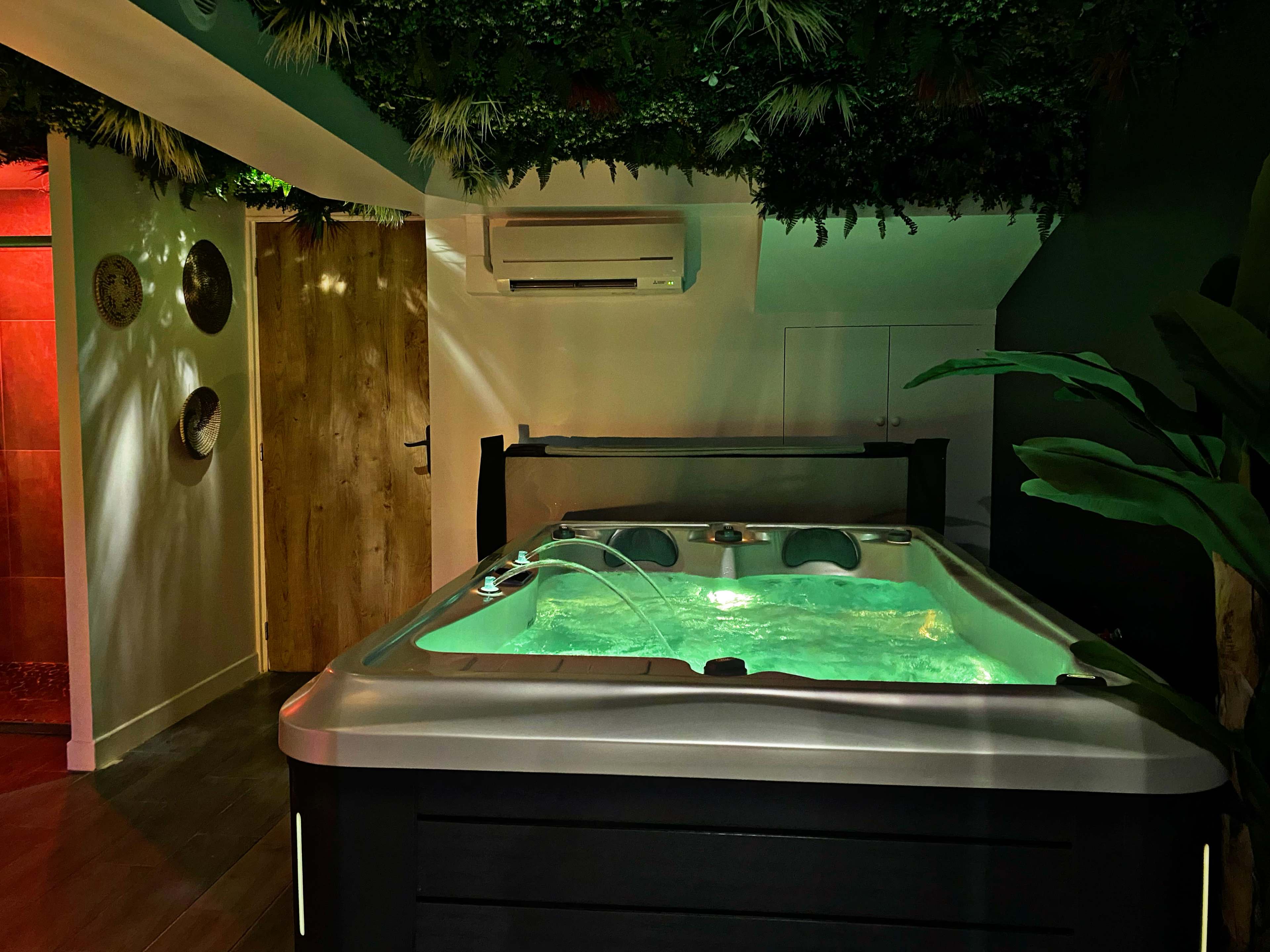 A modern indoor spa area featuring a hot tub with green lighting, surrounded by lush greenery and wooden accents.