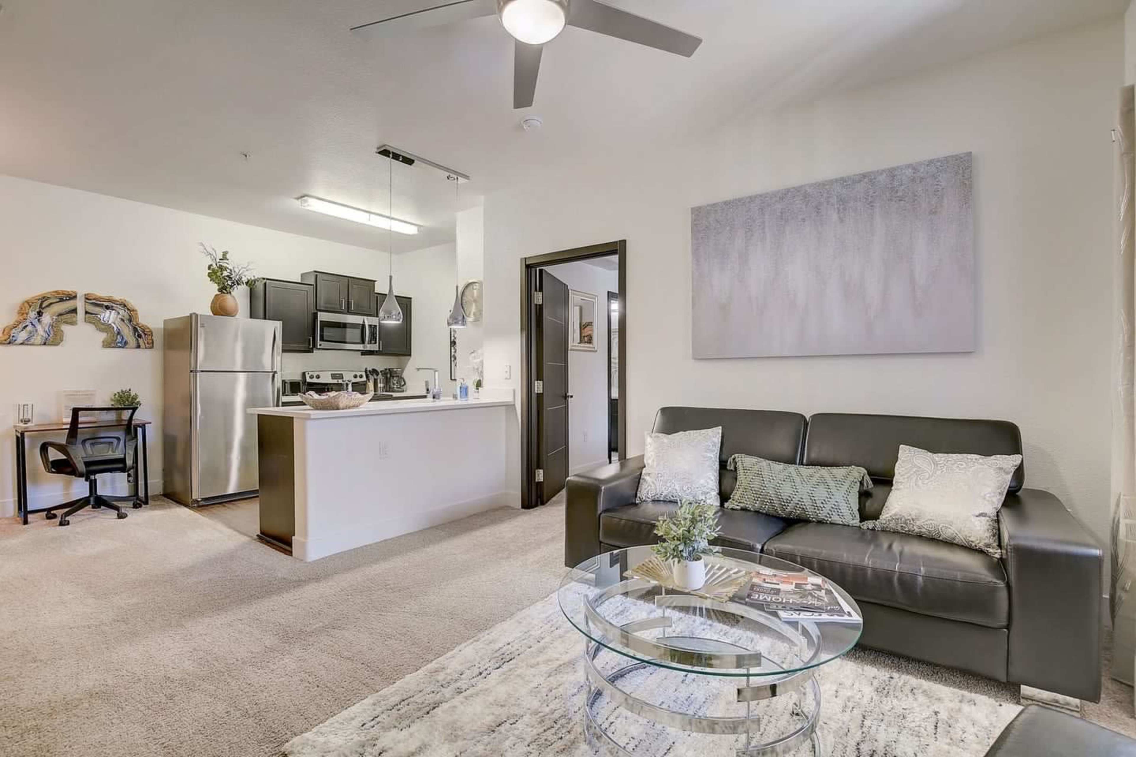 A modern living room with a black sofa, a glass coffee table, and a kitchenette in the background.