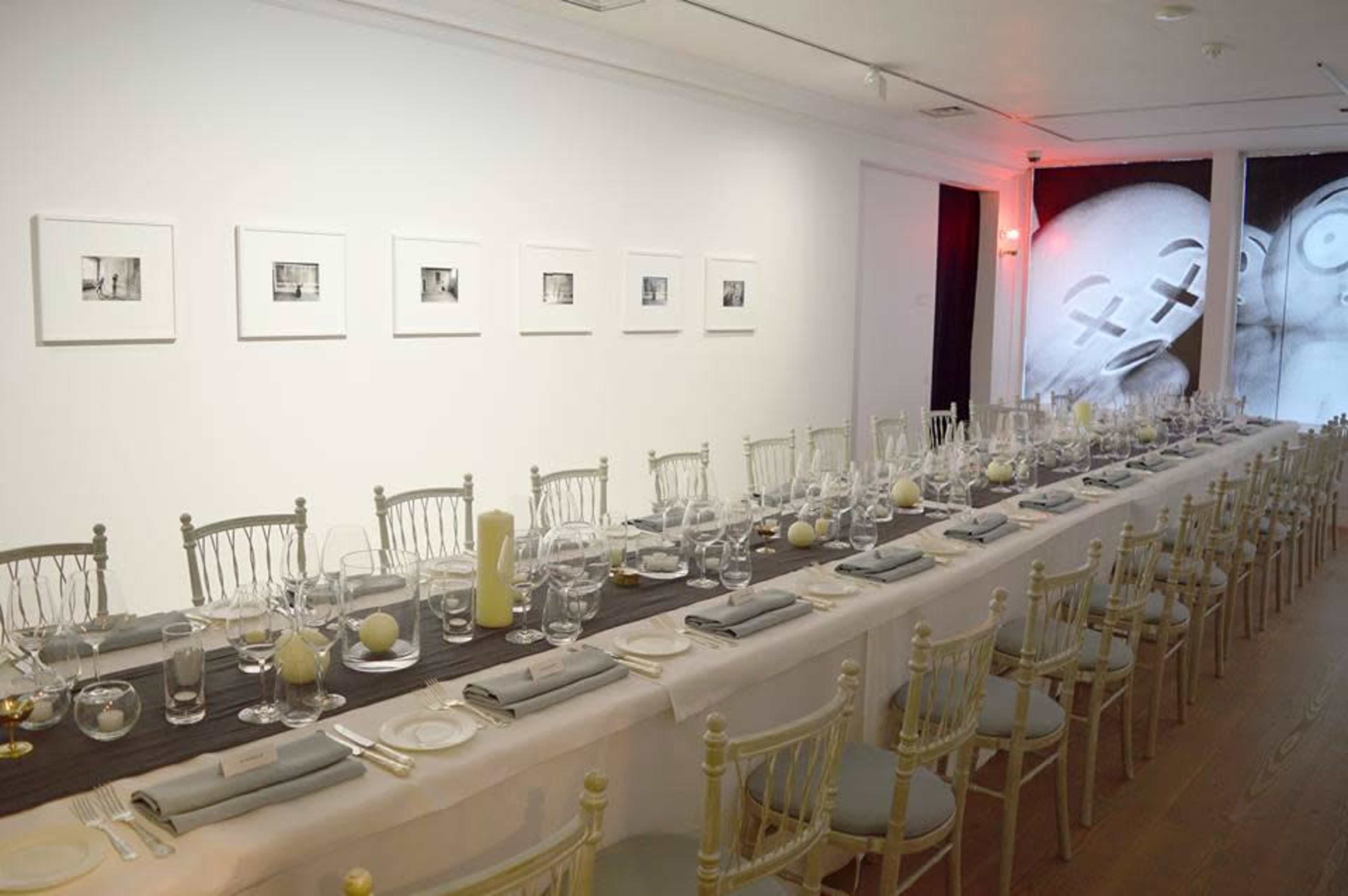 A long, elegantly set dining table is placed in a bright room, flanked by framed black-and-white photographs on the wall and a large abstract artwork in the background.