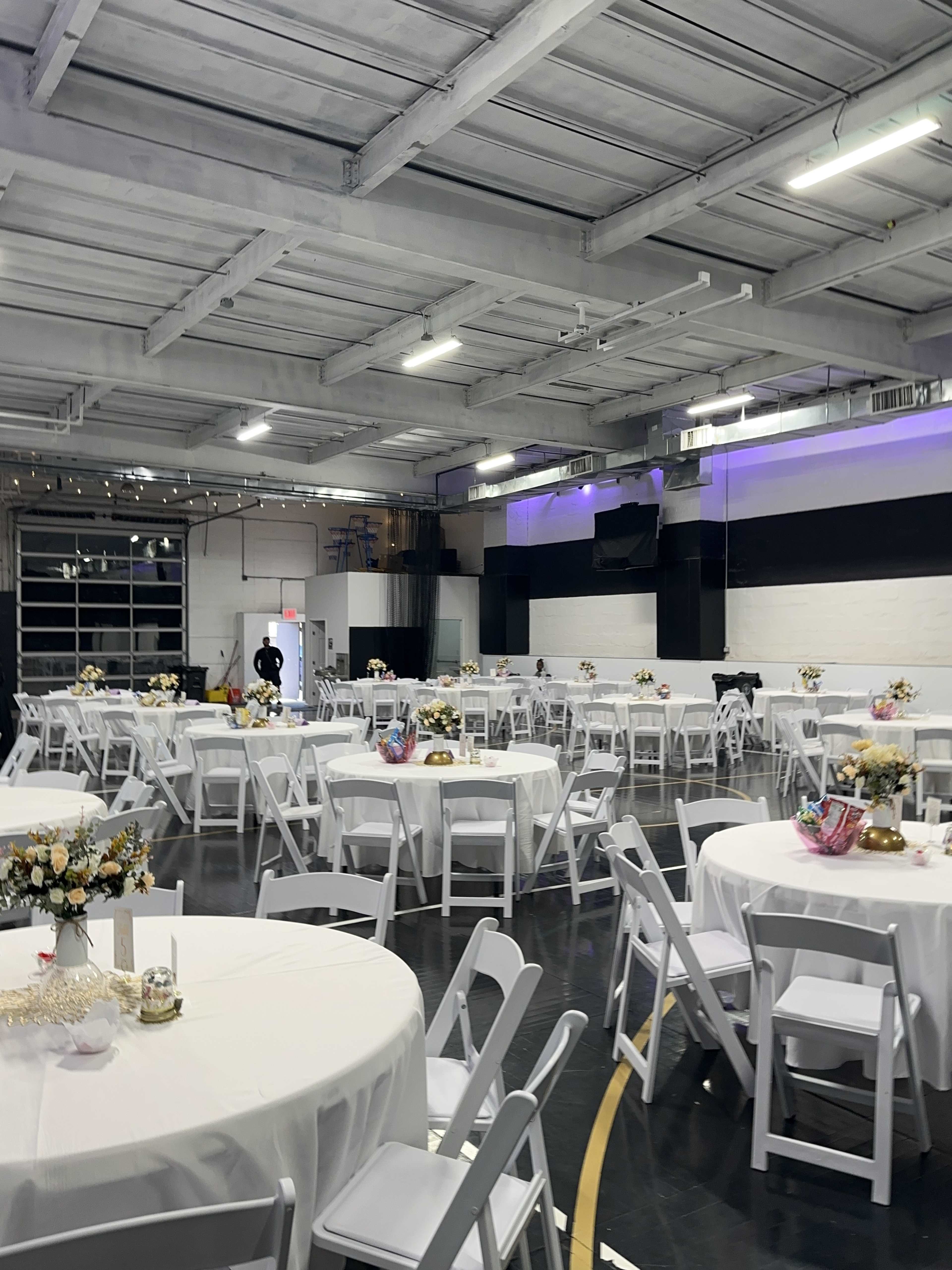 The image shows a spacious indoor venue decorated with round tables covered in white tablecloths, each featuring floral centerpieces.