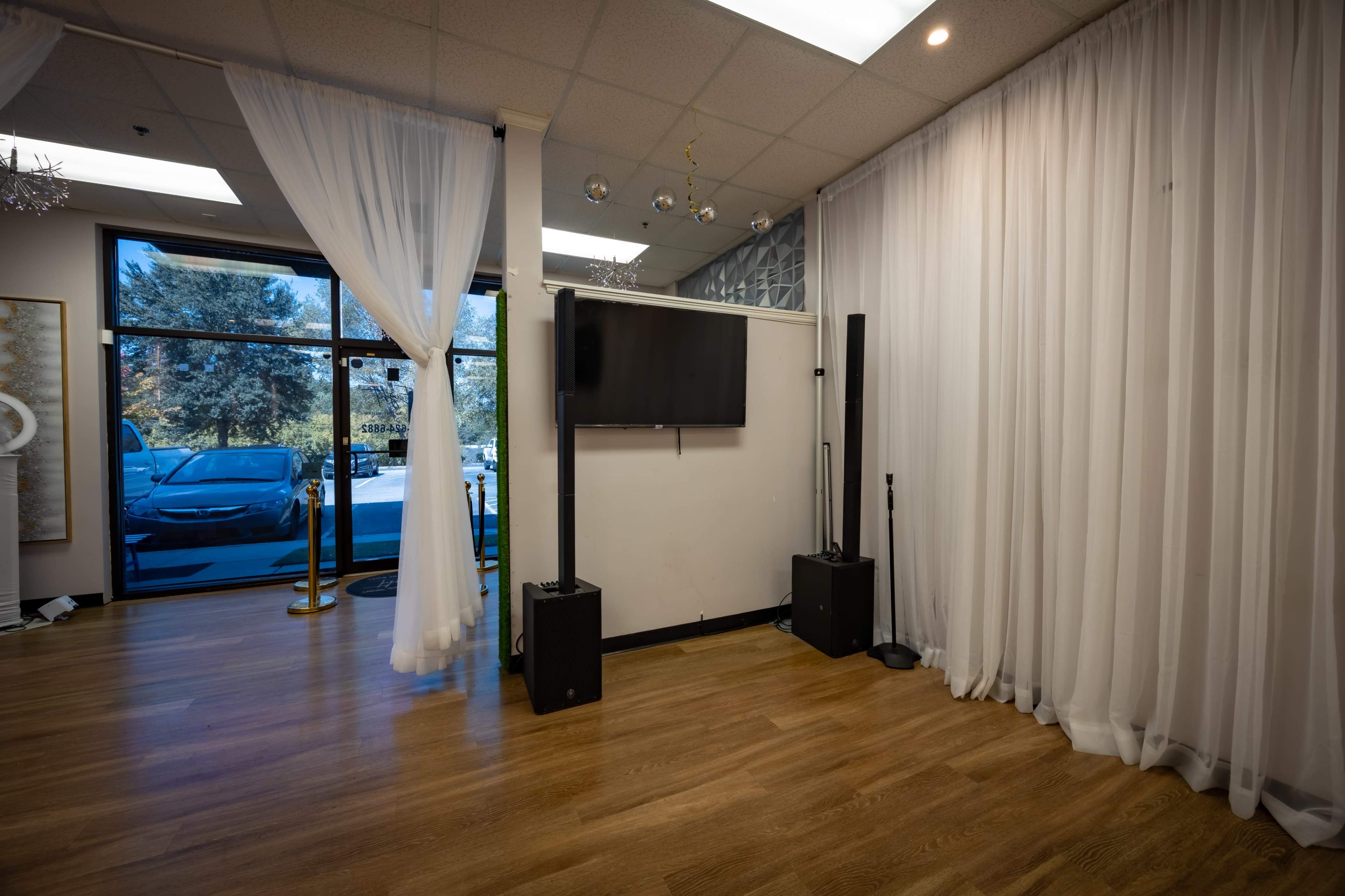 The image shows an interior space with wooden flooring, a wall-mounted television, and audio equipment, alongside sheer curtains and large windows.