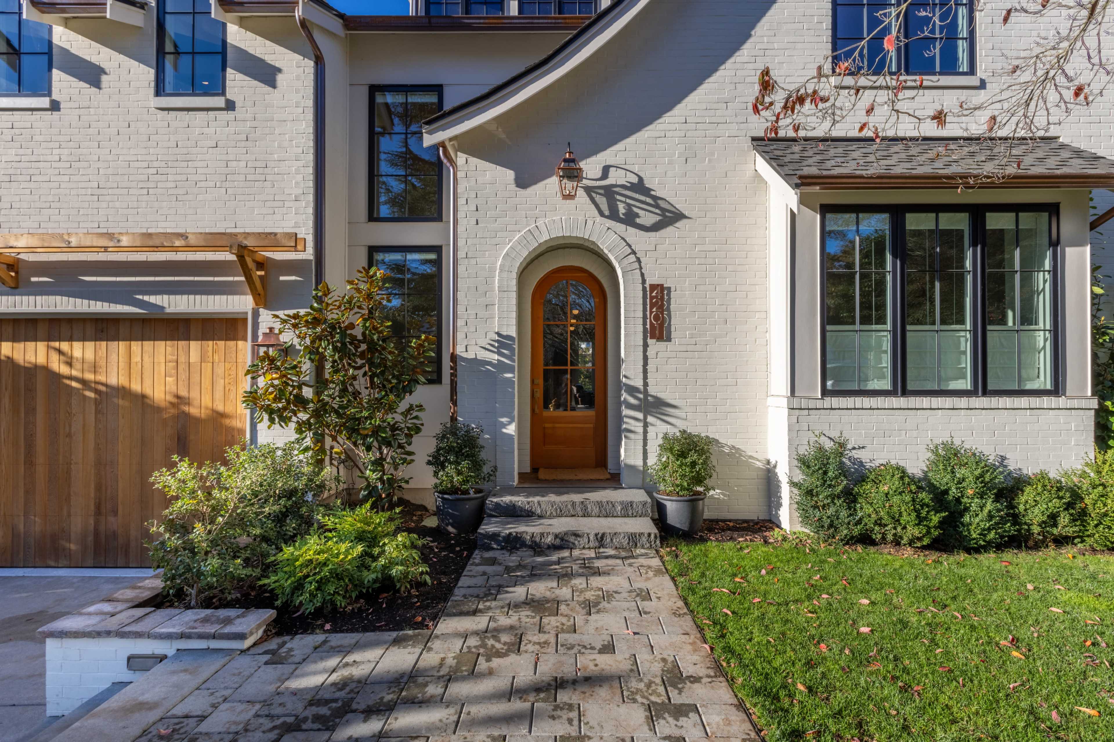 The image shows a well-maintained front entrance of a house with a wooden door, surrounded by greenery and a stone walkway leading to it.