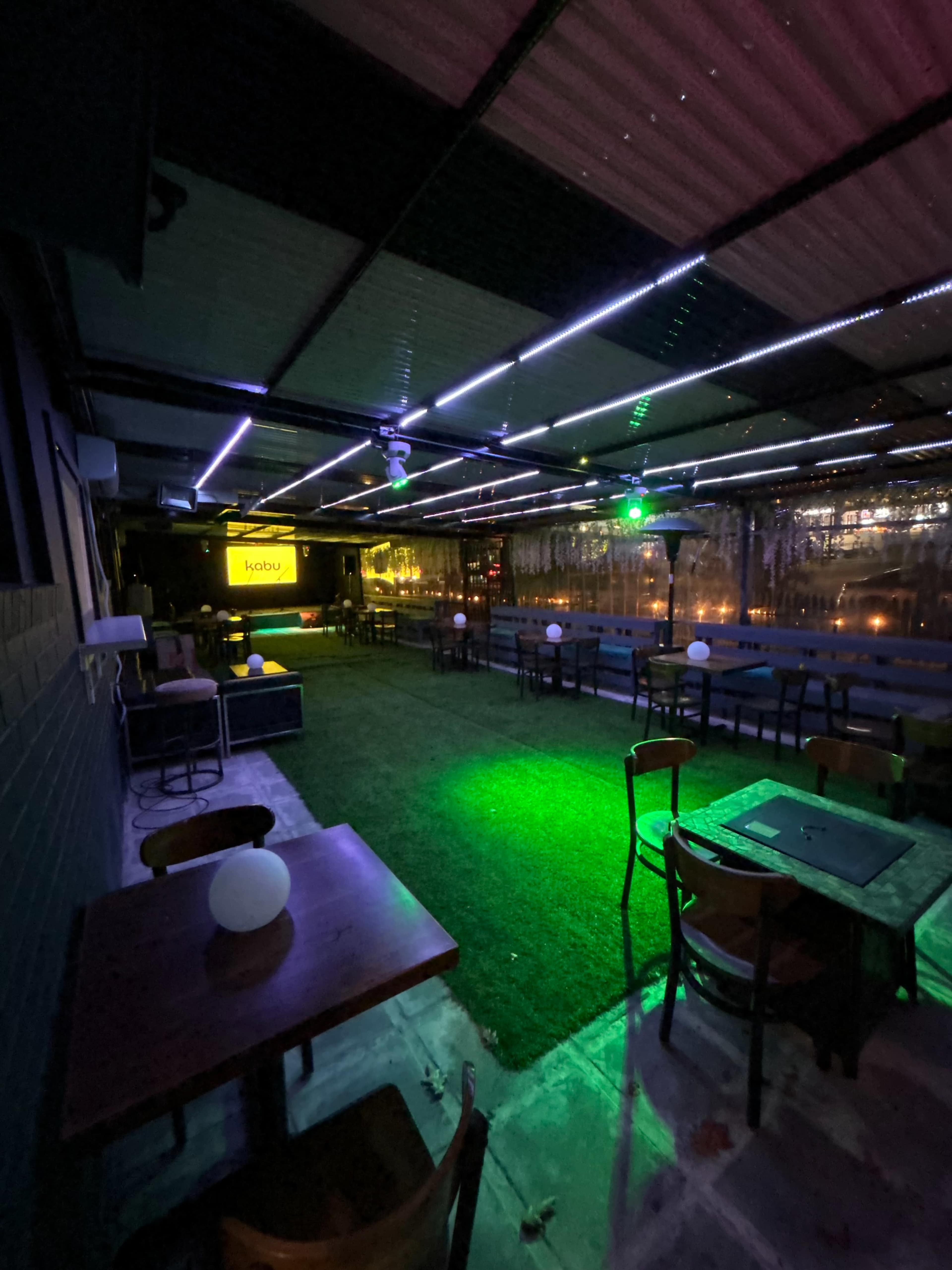 The image shows a dimly lit outdoor venue with green lighting, sparse seating, and a large screen displaying a logo in the background.