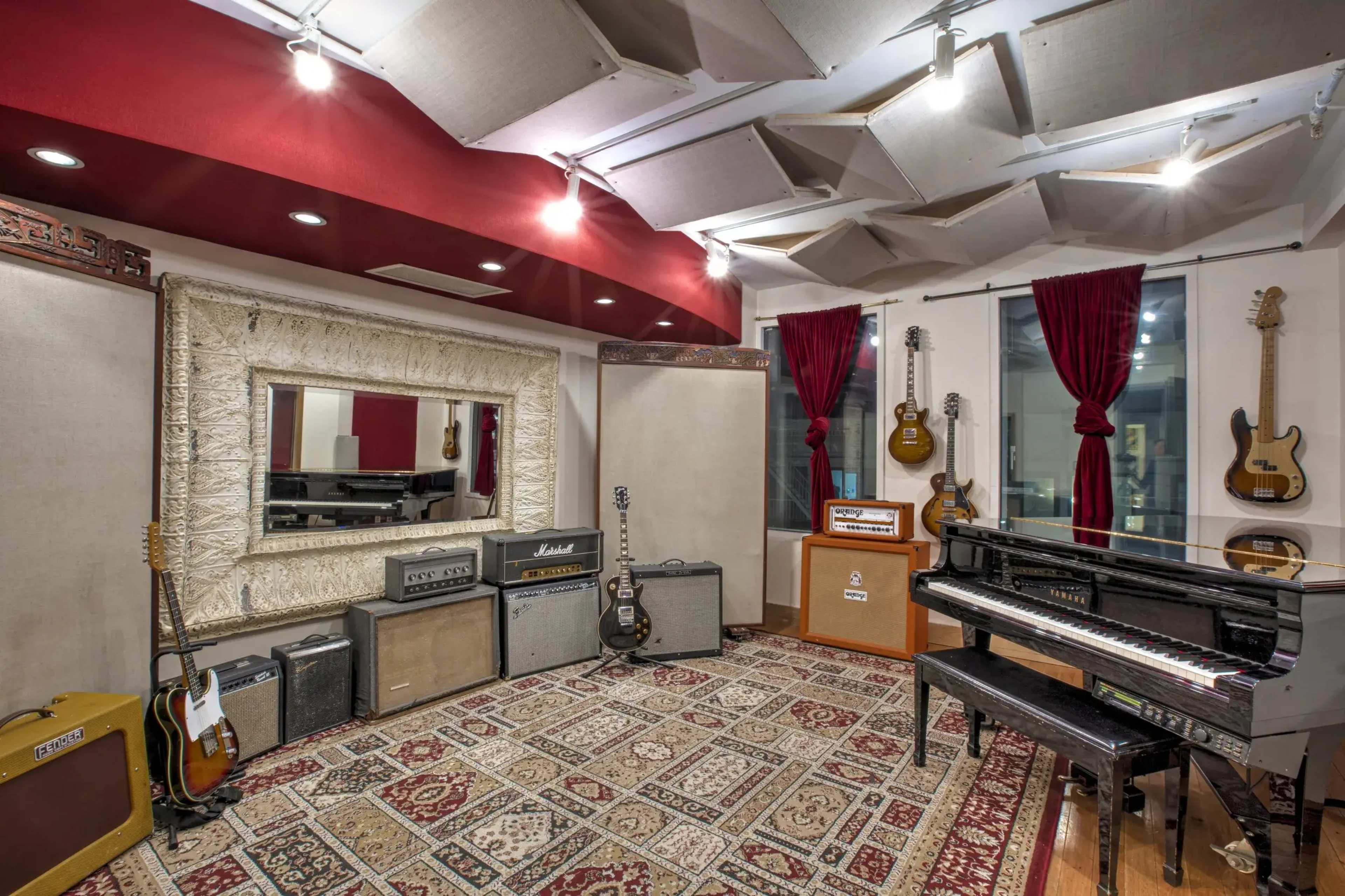The image shows a music studio featuring various amplifiers, a grand piano, and guitars displayed on the walls, with soundproofing panels and decorative curtains.