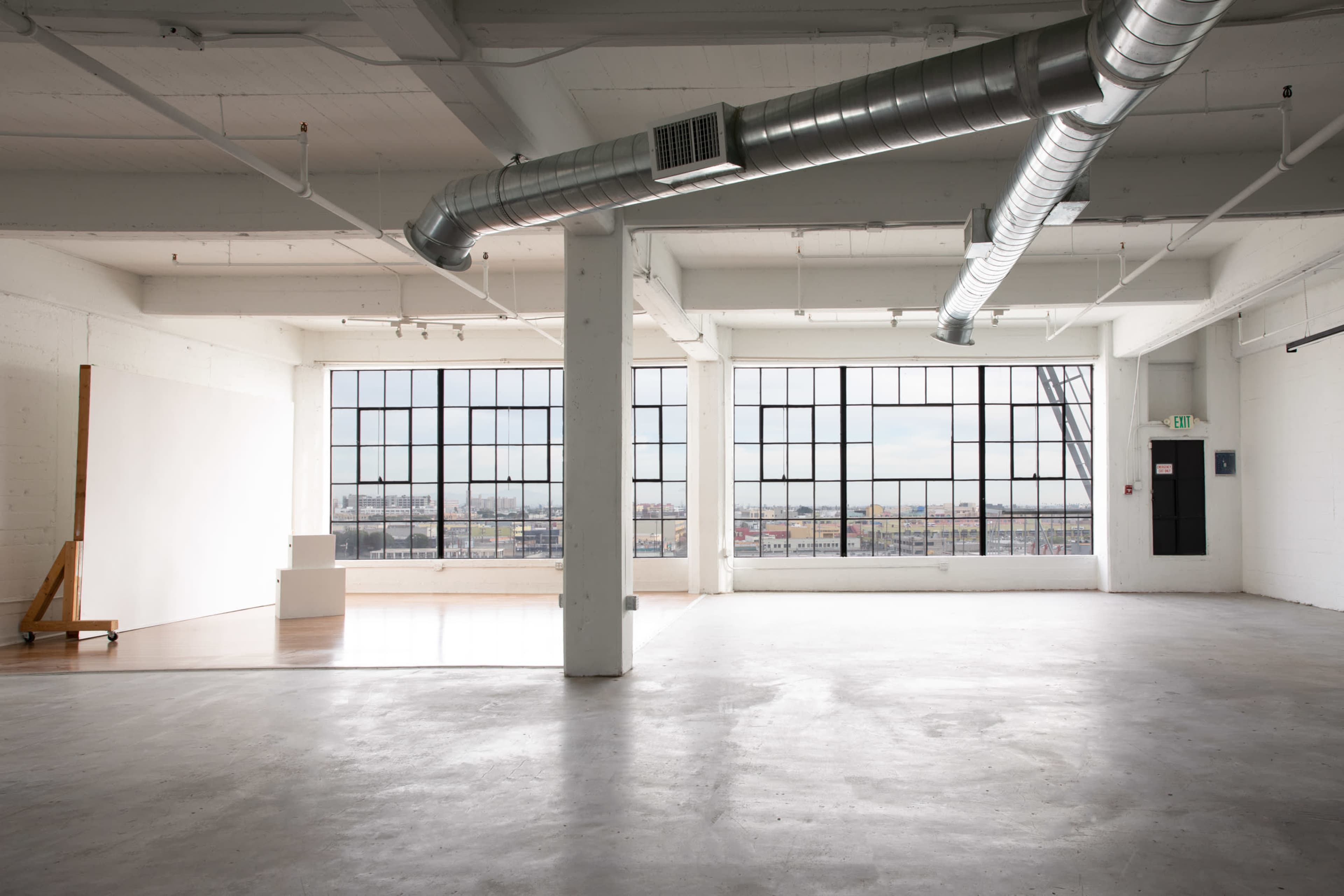 The image shows a spacious, industrial-style room with large windows and concrete floors, featuring minimal furnishings.
